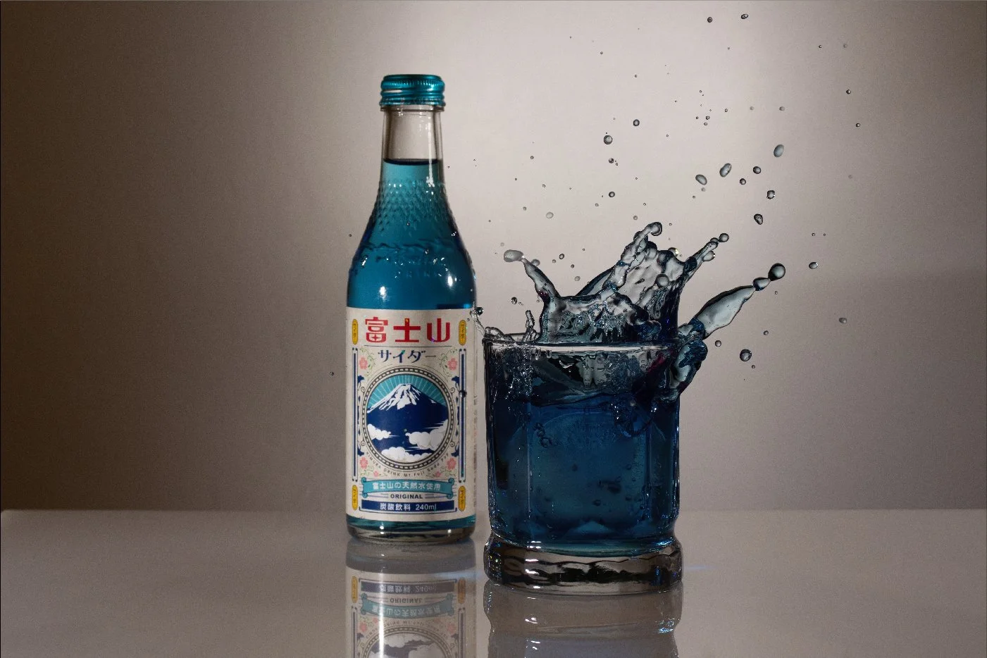 A glass of blue soda with ice and a bottle of Fuji Mountain soda next to it, with bubbles splashing in the glass.