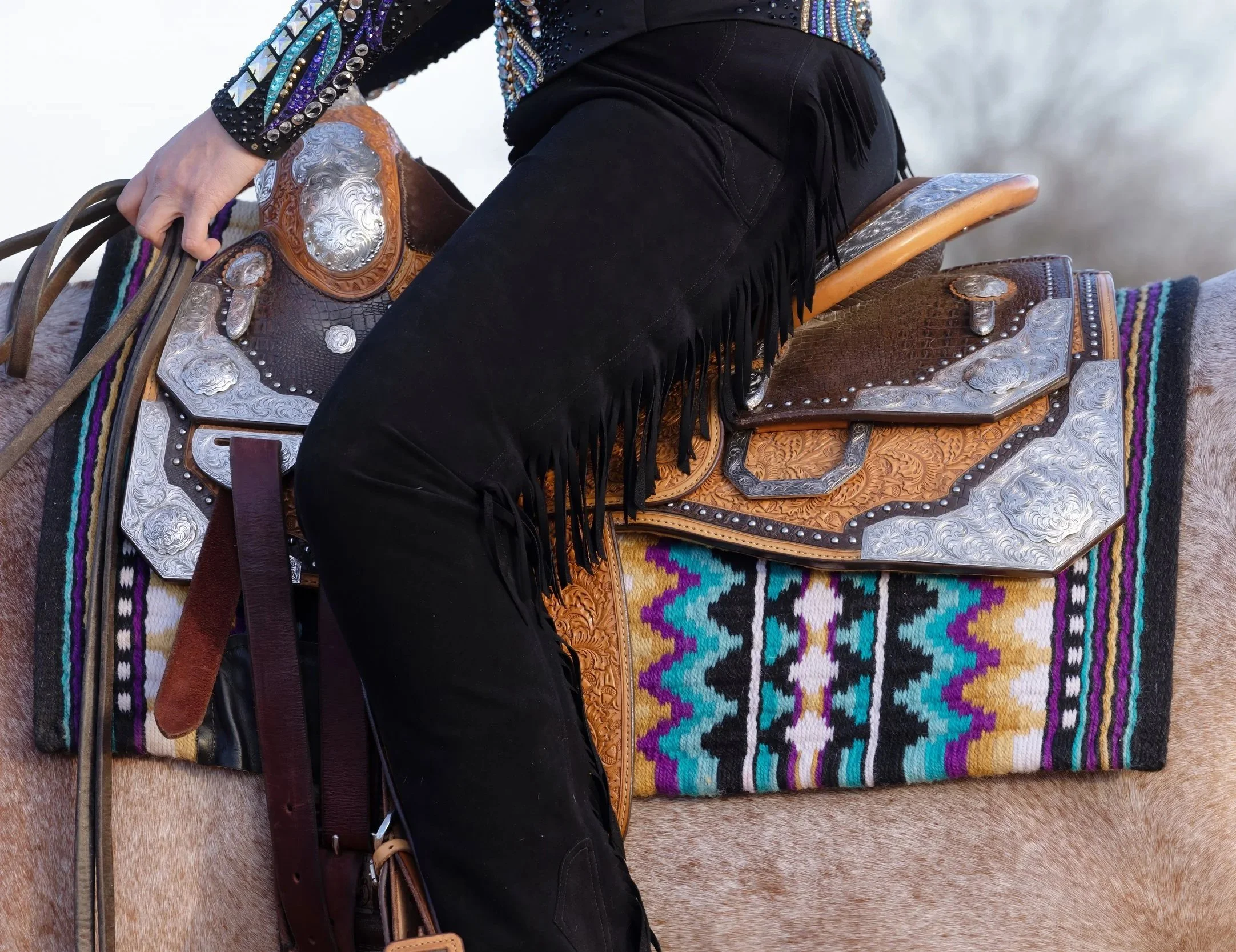Close-up of a person dressed in Western attire on a saddle, with focus on their black fringed shirt, black pants, and part of the saddle decorated with silver and leather details, set against a blurred outdoor background.