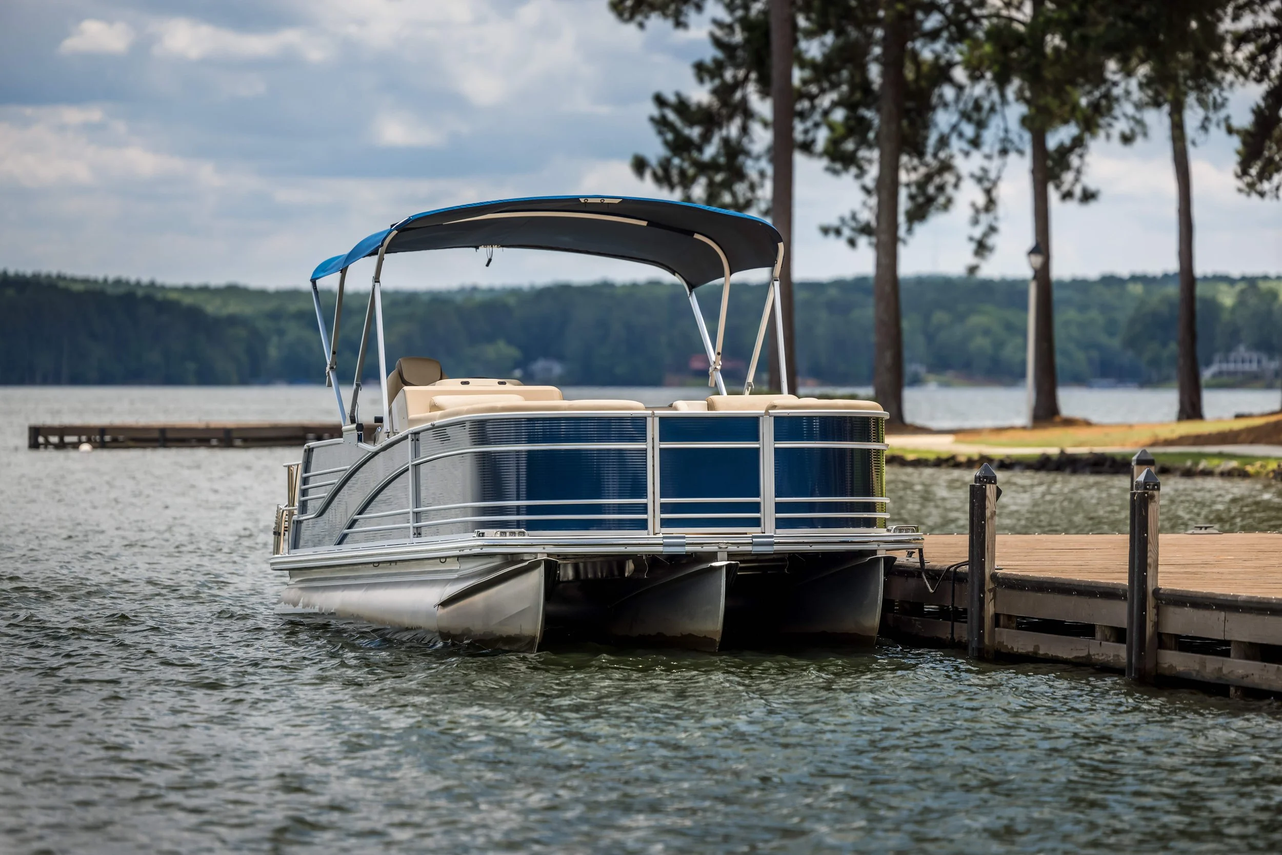 A pontoon boat docked at a wooden pier on a lake, with trees in the background and partly cloudy sky.