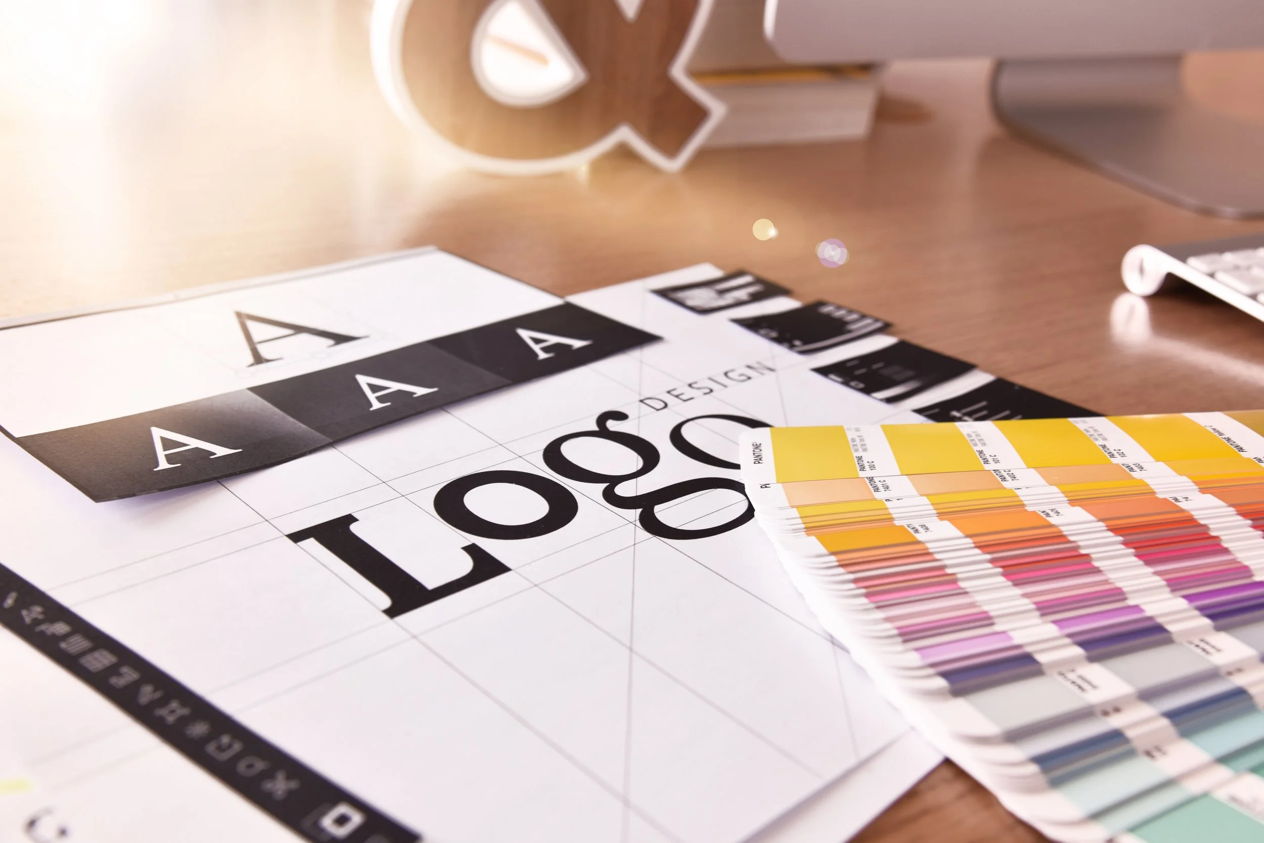 Design and color palette swatches, letters 'A' and 'O' on a printed sheet, yellow and orange color samples, a white notebook in the background, and a wooden desk surface.