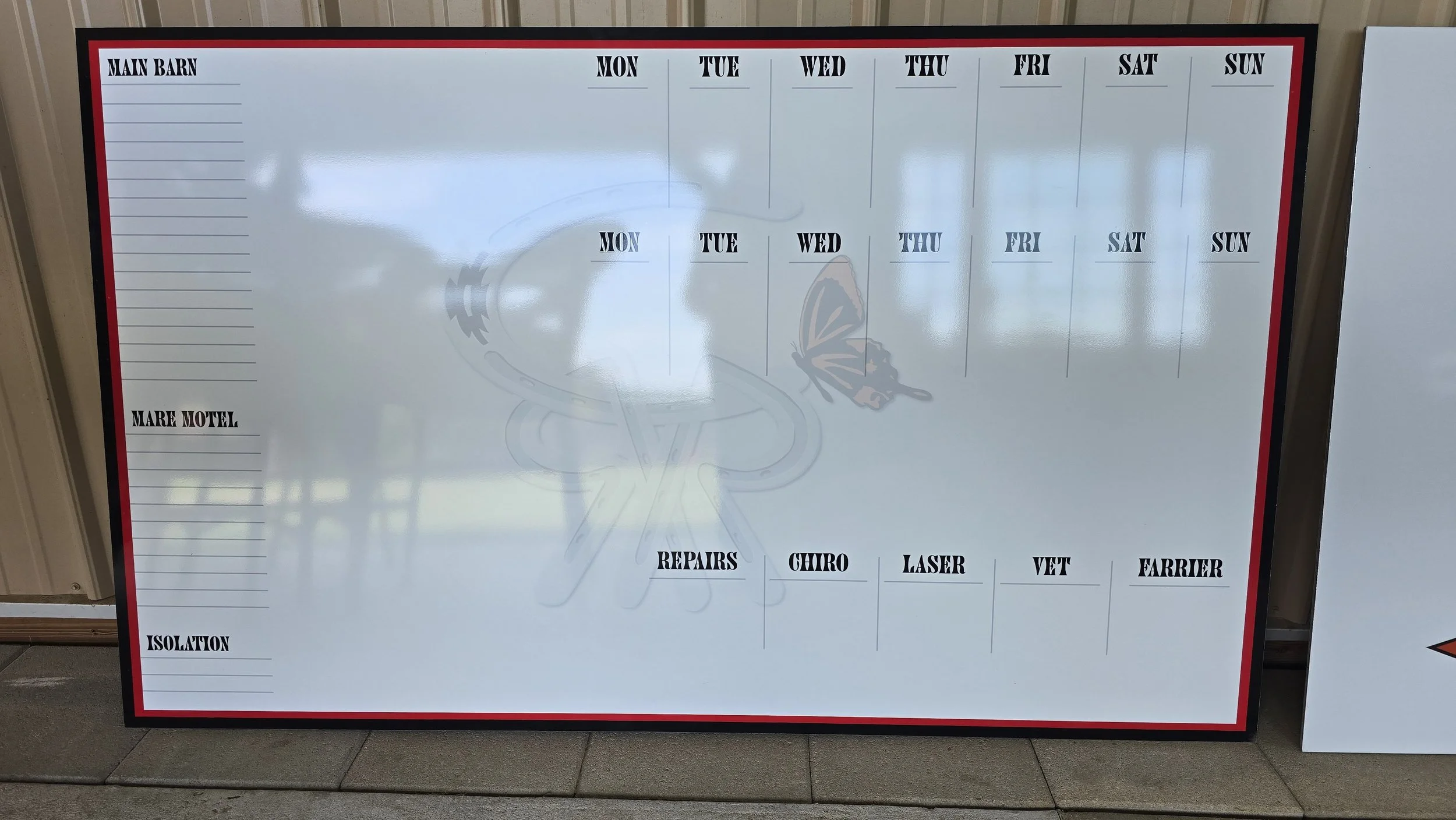 A large whiteboard with a weekly schedule, divided into days of the week, with sections labeled for main barn, mare motel, repairs, chiros, laser, vet, farrier, and isolation. The background has a faint image of horseshoes and a butterfly.