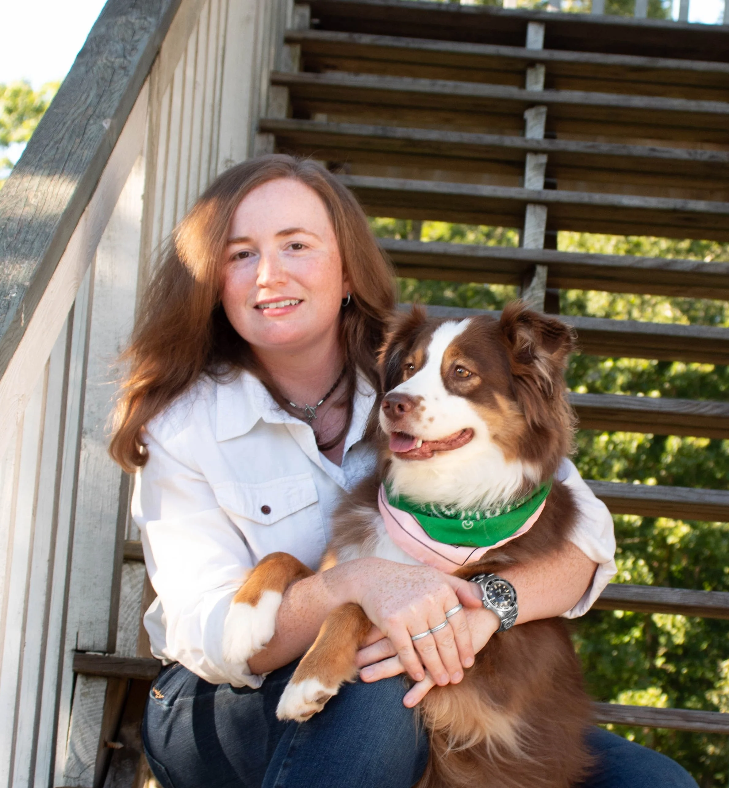 A woman with long brown hair, wearing a white shirt and a watch, holding a brown and white dog with a green bandana outdoors on a wooden staircase.