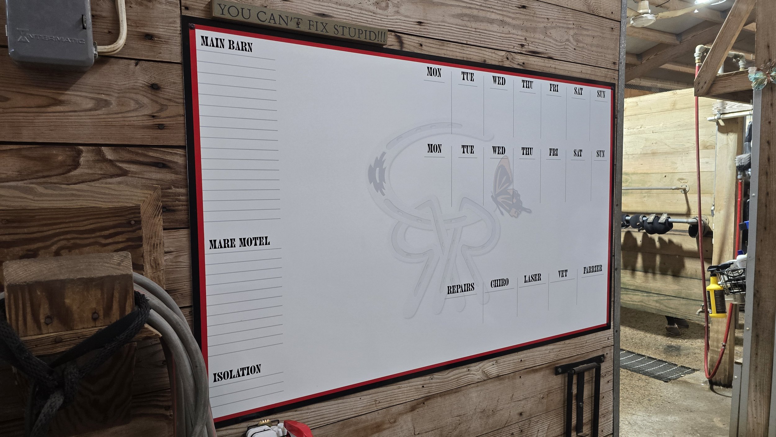 Whiteboard calendar or planner with weekly sections for Monday to Sunday, located in a workshop or barn area with wooden wall panels, pipes, tools, and equipment nearby.