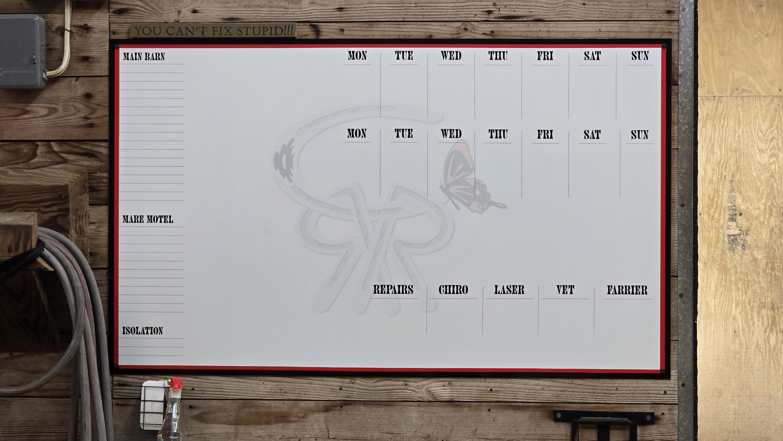 A white board with a weekly schedule layout on a wooden wall. The board has sections labeled for main barn, mare motel, repairs, chiro, laser, vet, farrier, and isolation, with days of the week from Monday to Sunday. 