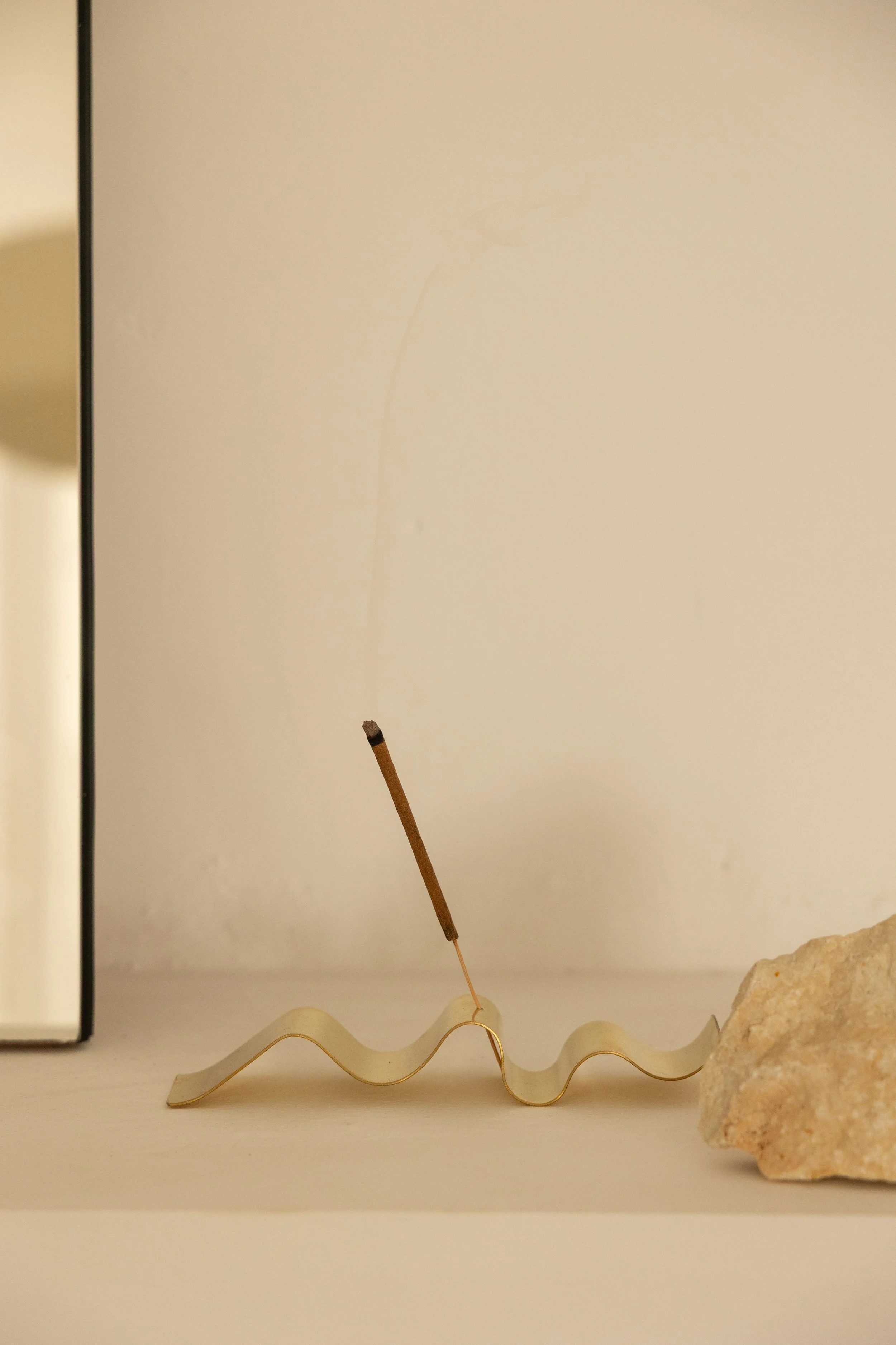 A decorative arrangement on a shelf with a wavy gold-colored metal piece, a small rock, a thin incense stick, and part of a framed picture in the background.