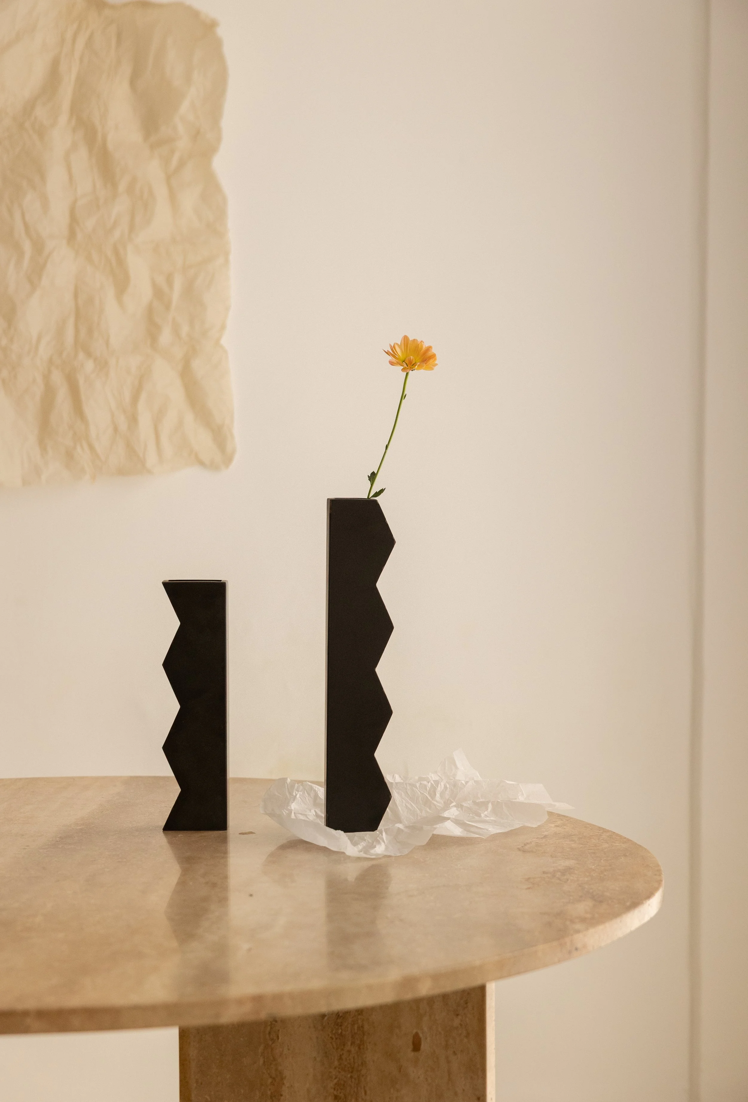 A small orange flower in a black geometric vase on a beige table, with textured white paper in the background.