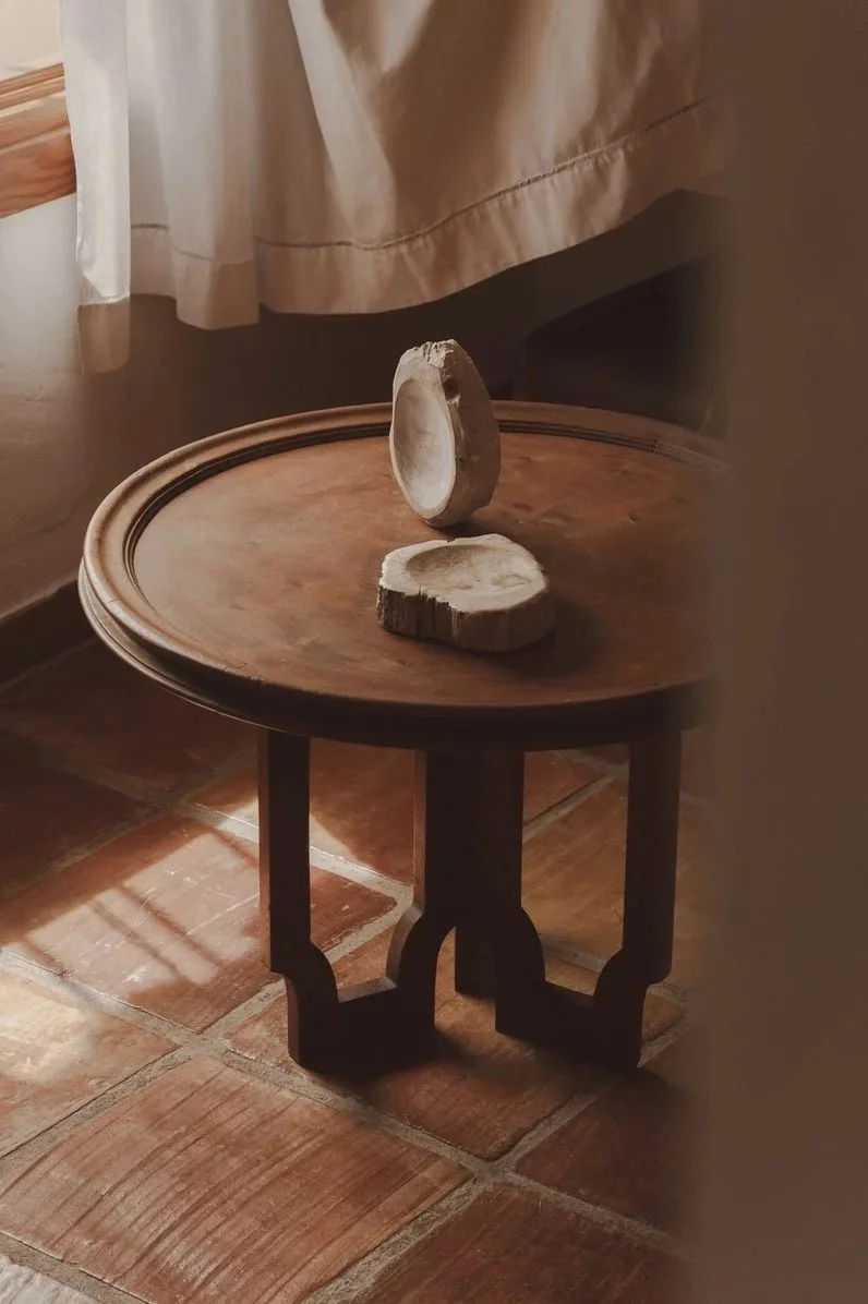A small wooden table with a round top, on which a sculpture of two wooden slices with concrete in the middle is placed, with a white curtain in the background.