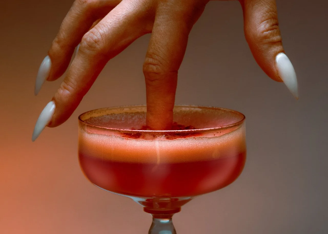 Person with long white nails dips finger into a pink cocktail drink with foam on top