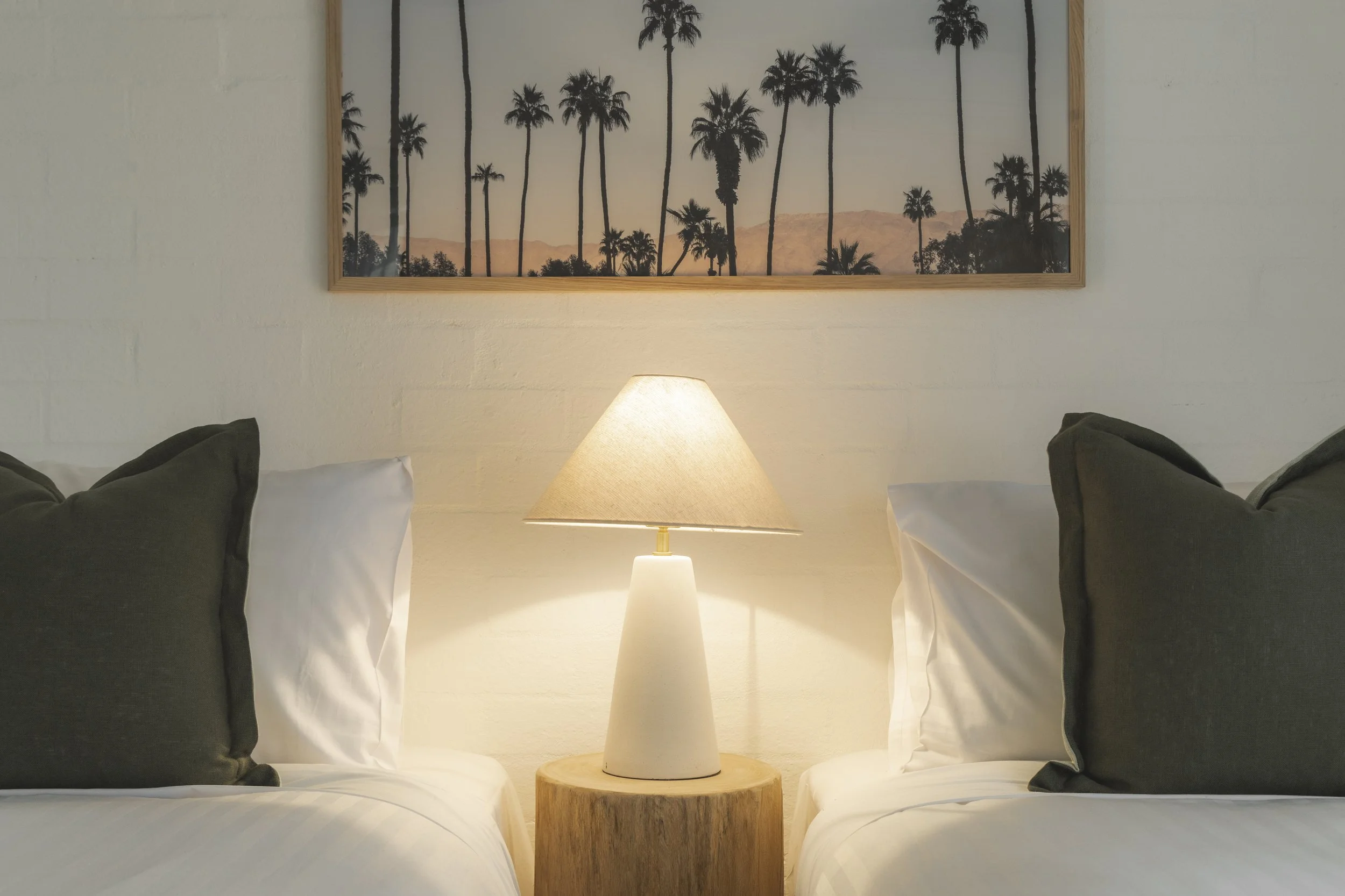 A bedroom with a nightstand, table lamp, framed picture of palm trees on the wall, and beds with pillows.