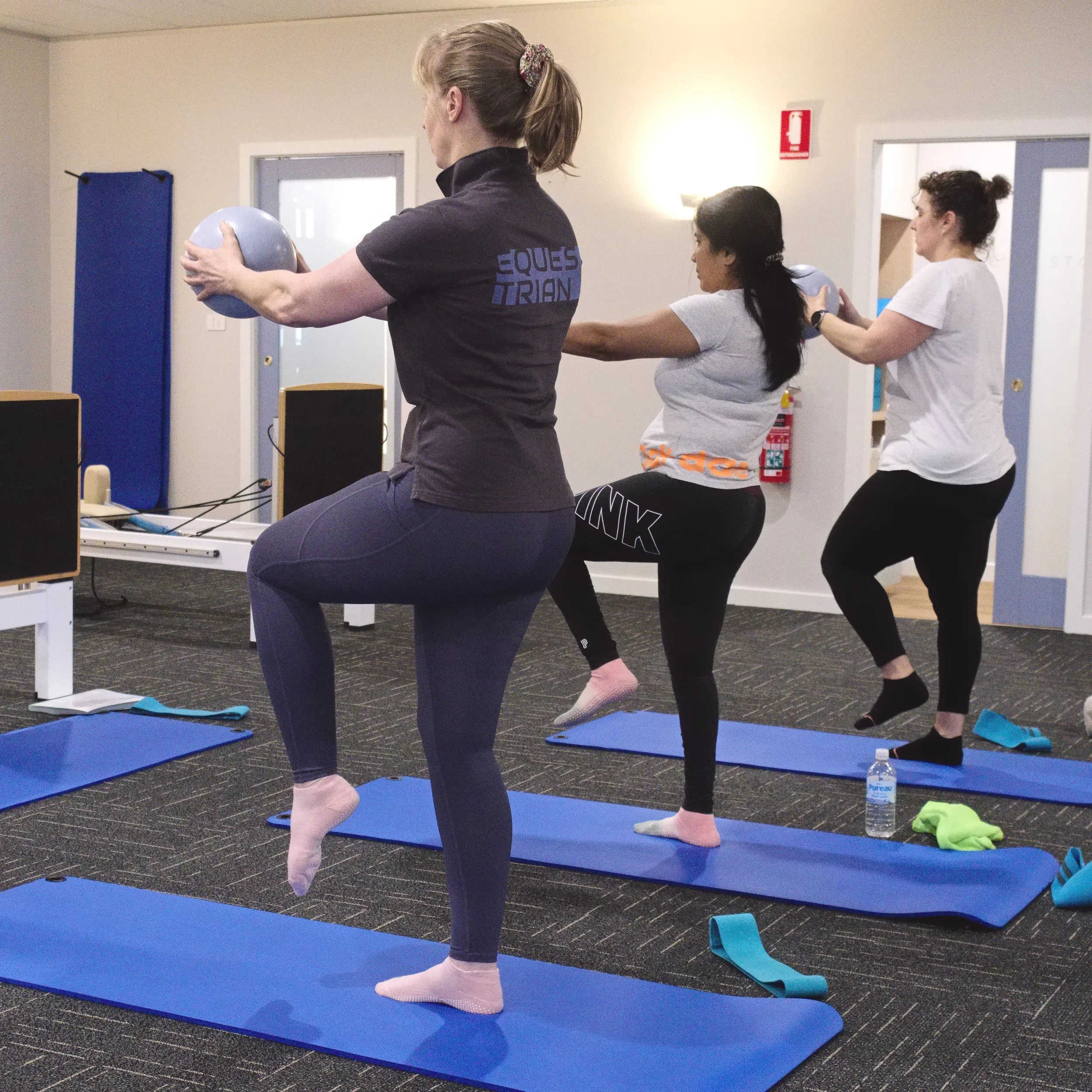Three women practicing fitness exercises on blue yoga mats in a gym, holding small exercise balls in front of them, with a water bottle and towel on the floor.