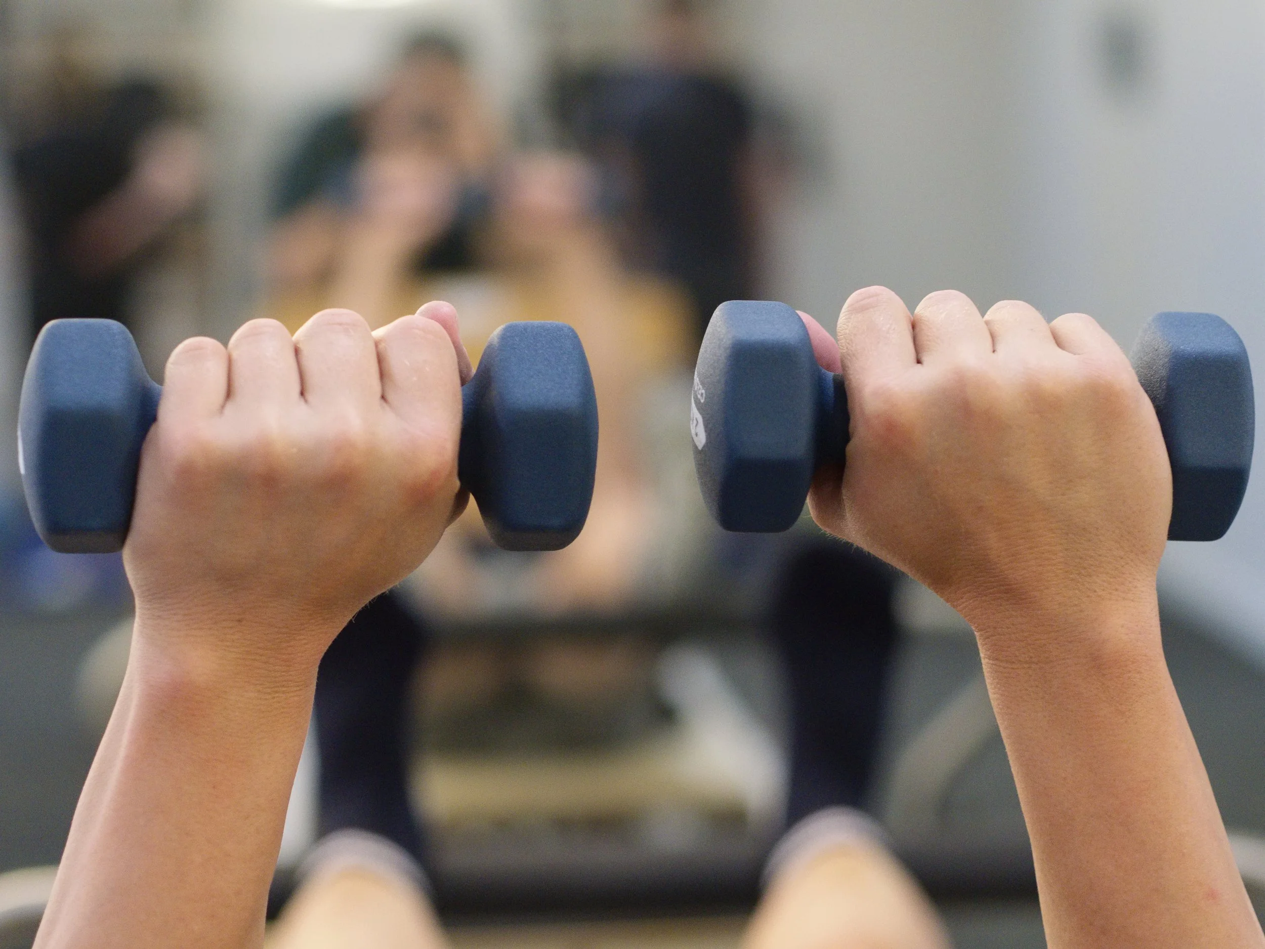 Hands gripping a pair of blue dumbbells, with a mirror showing students in workout attire in the background.