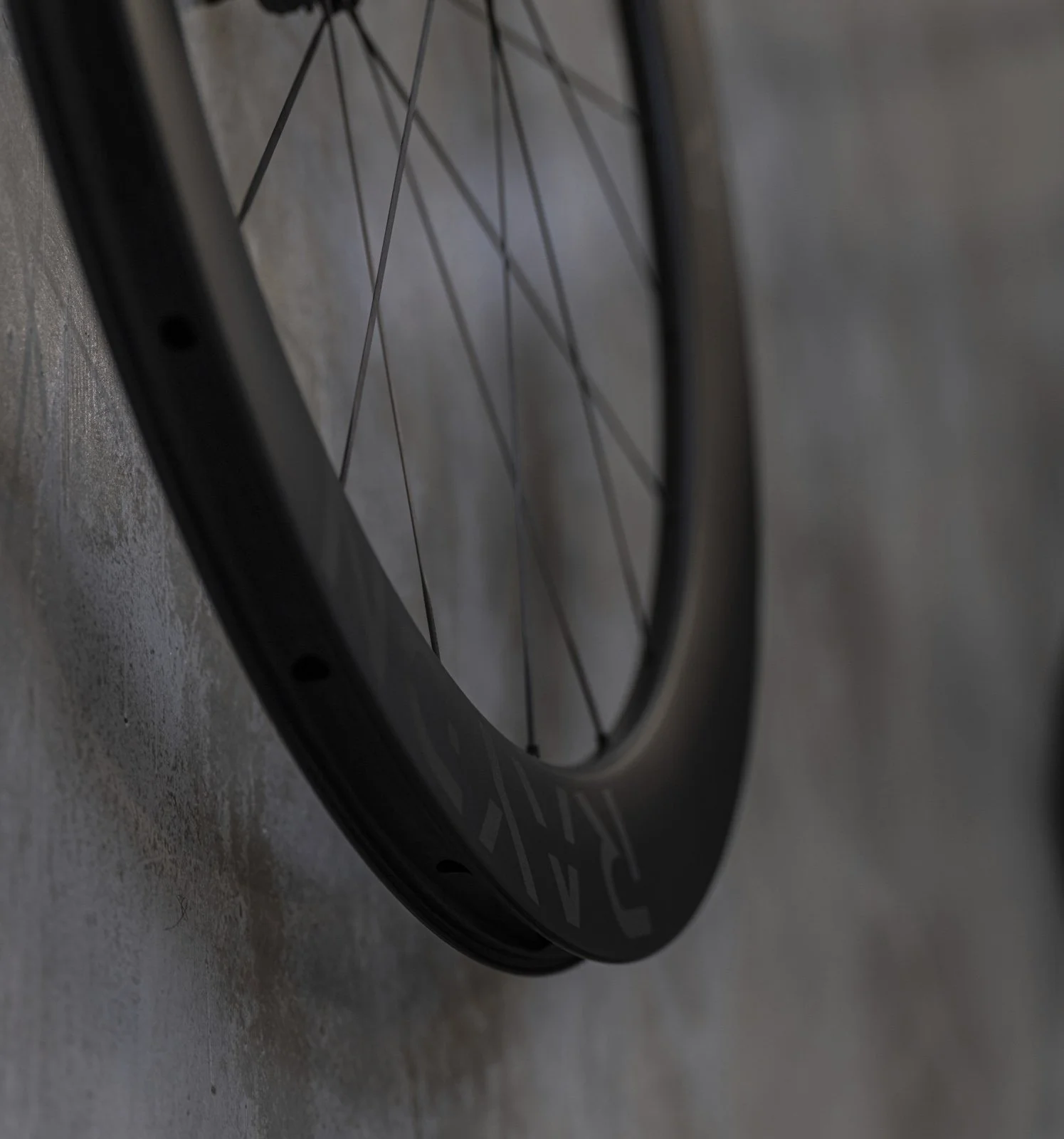 Close-up of a carbon bicycle wheel with spokes, lying against a textured concrete surface.