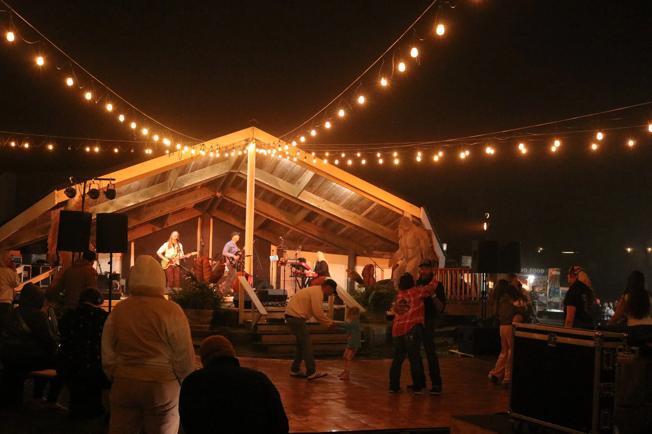 Night scene of an outdoor music event with a band performing on a wooden stage under a peaked roof, surrounded by string lights and an audience dancing and socializing.