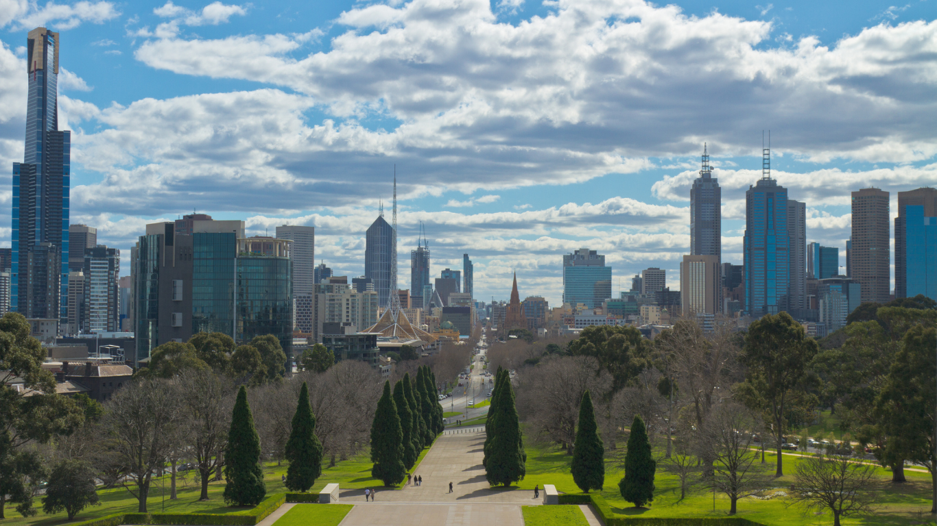 A city skyline with tall skyscrapers behind a park with trees and a pathway in the foreground, under a partly cloudy sky.
