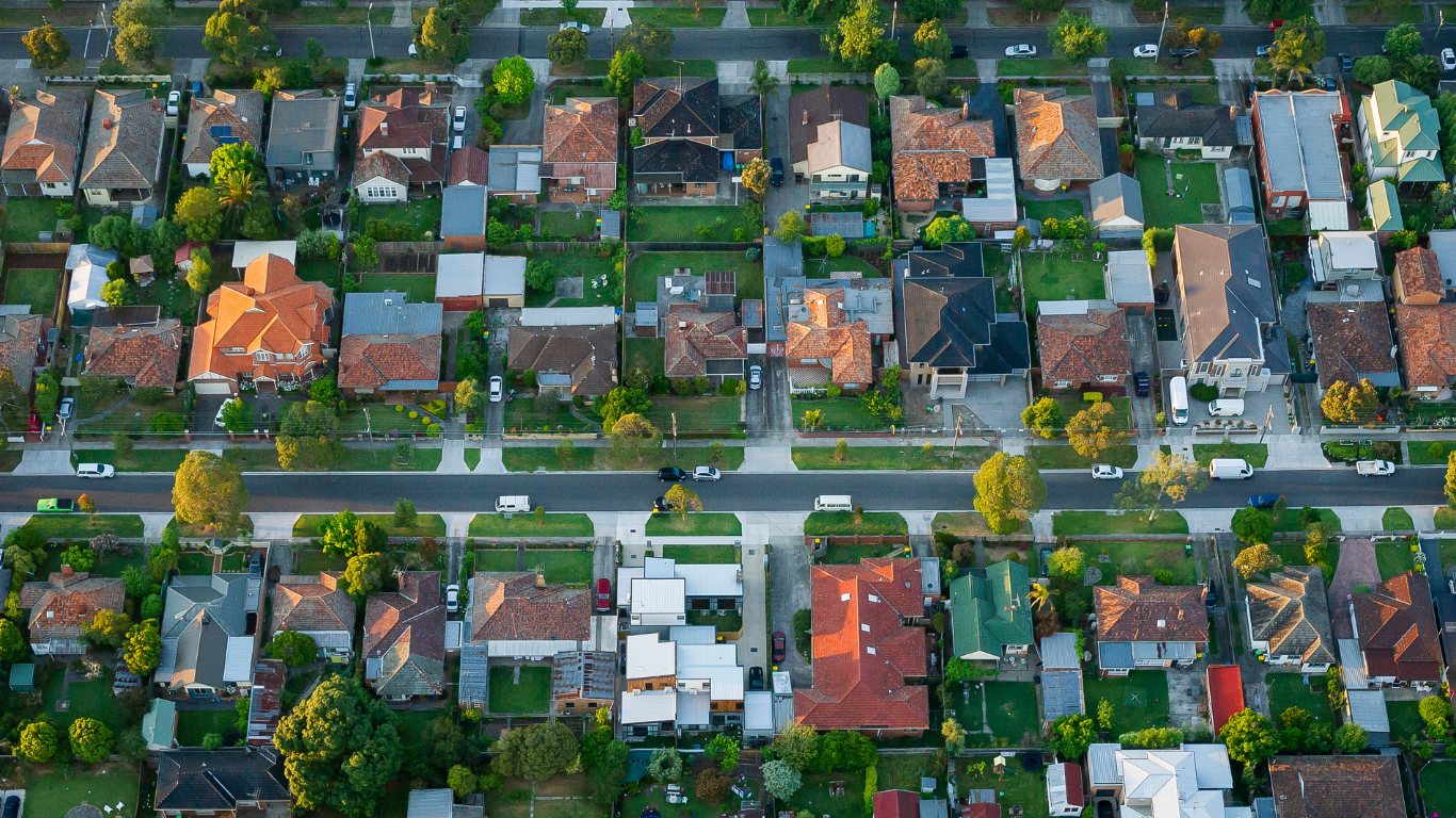 An aerial view of a suburban neighborhood with houses, trees, and parked cars along streets.