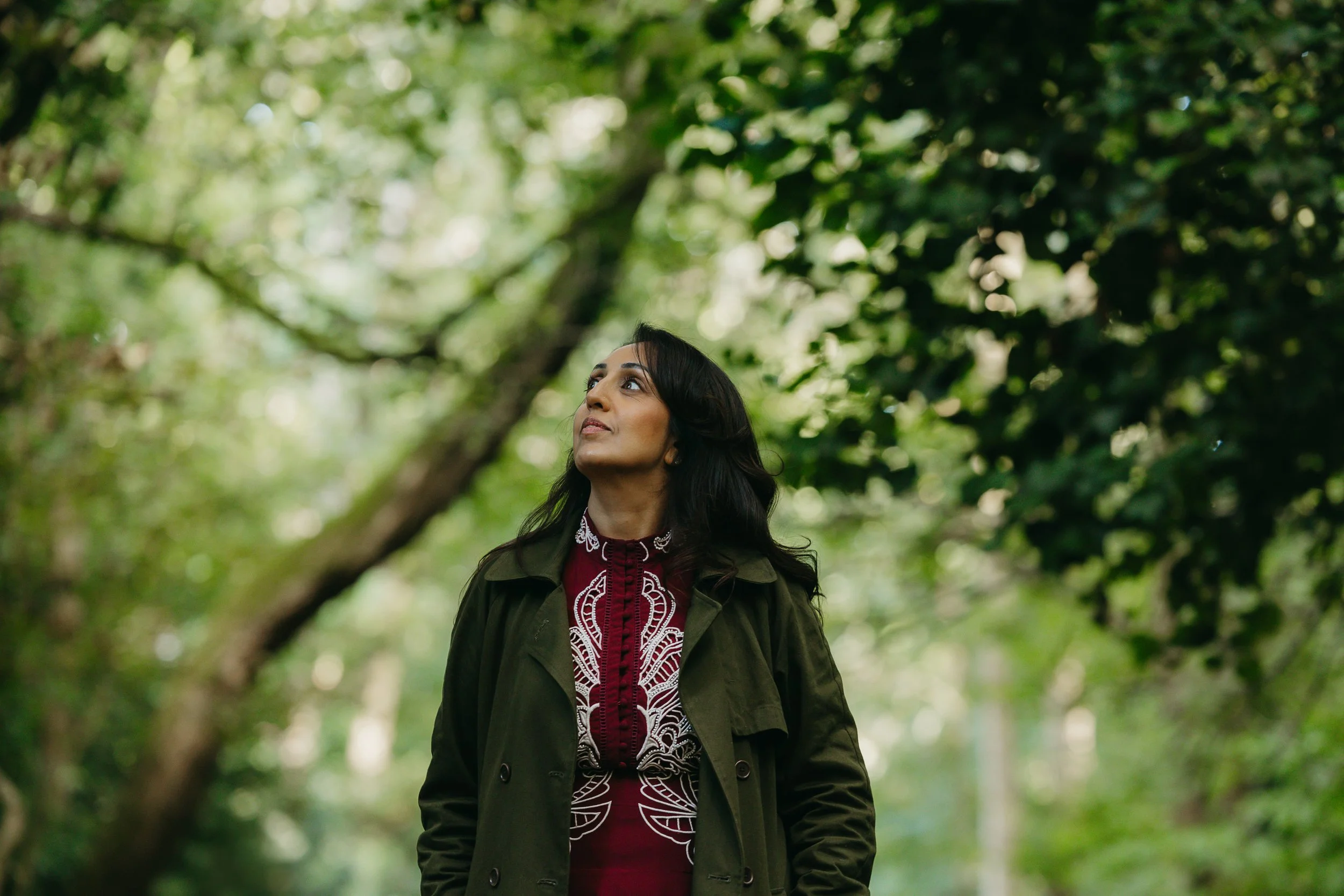 A woman with dark hair standing in a lush green forest, looking upward with a serene expression.