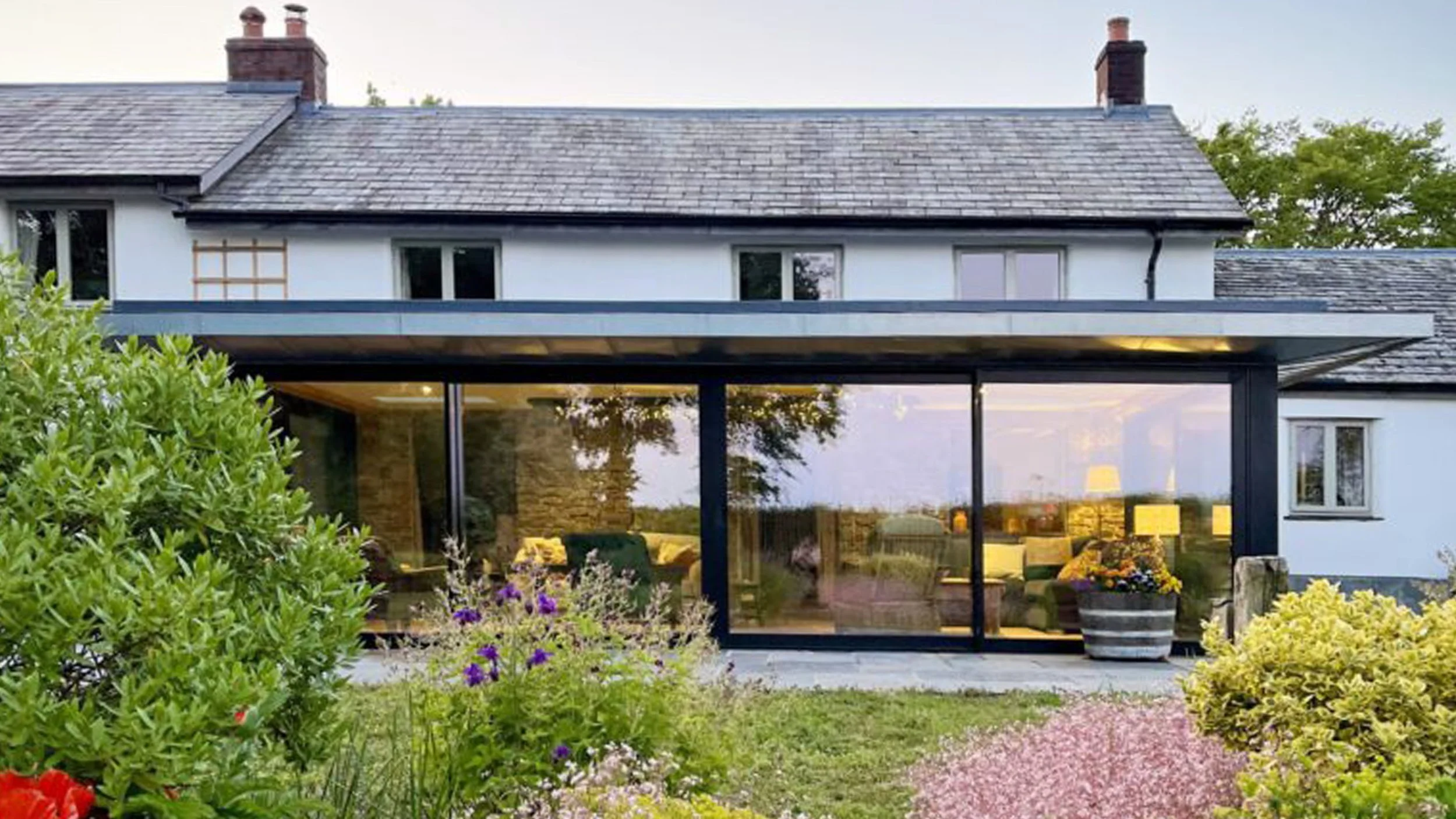 L&M Architects Ltd — A modern two-story house with large glass windows and a garden in front, featuring various colorful flowers and green bushes.