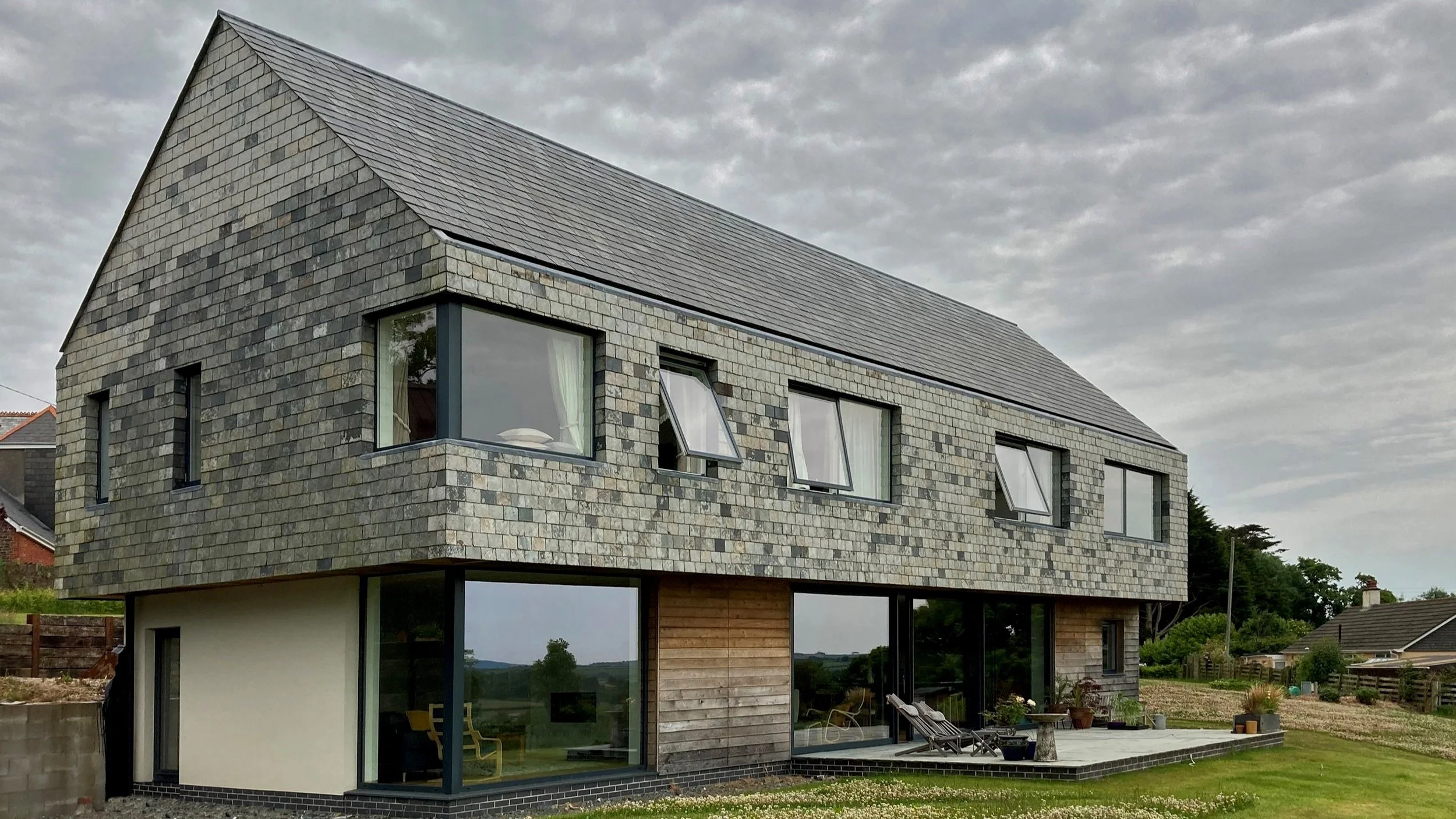 L&M Architects Ltd — Modern multi-story house with stone and wood exterior, large windows, and a sloped roof, situated in a suburban area under a cloudy sky.