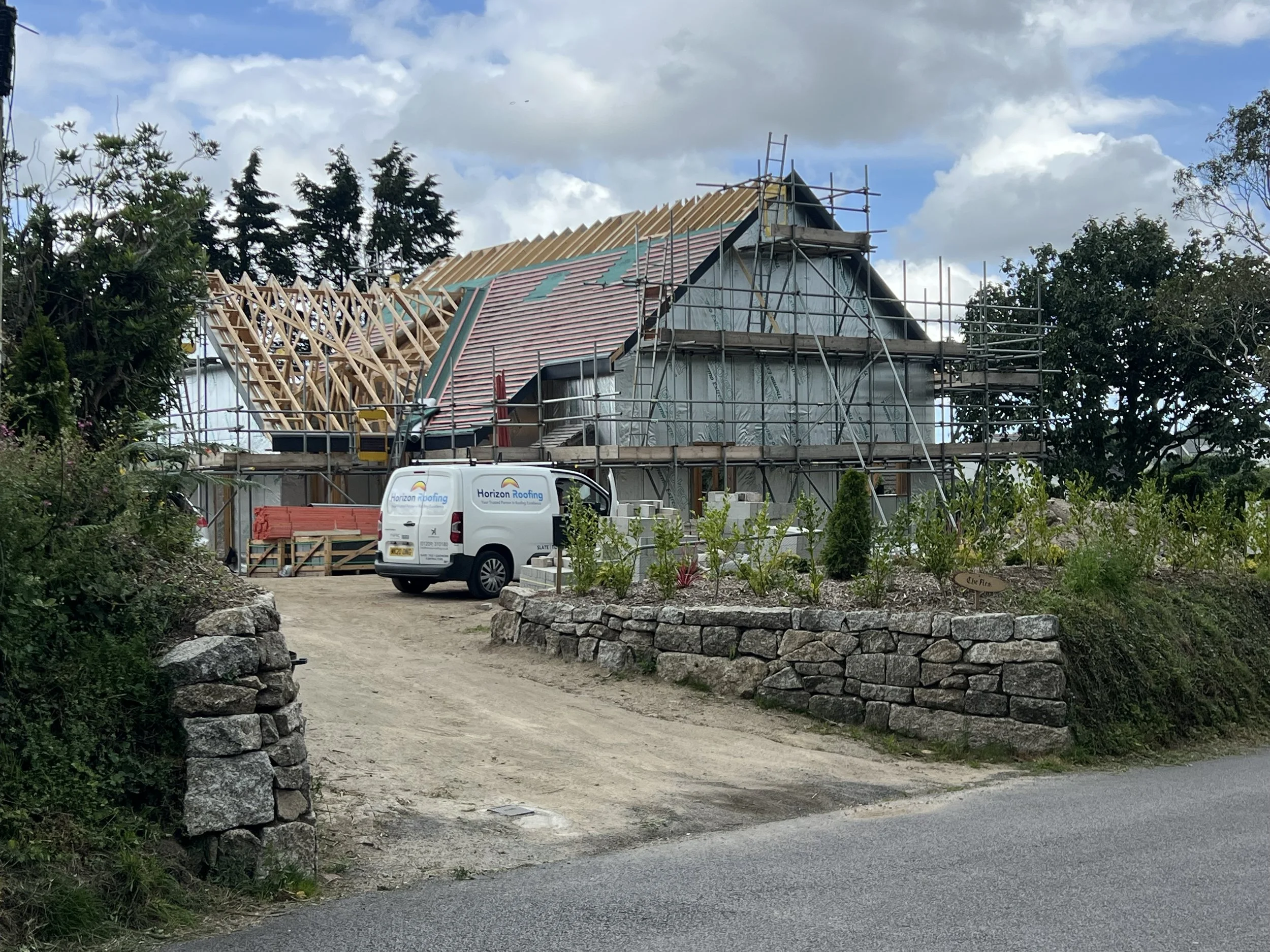 A house under construction with scaffolding, a partially finished roof, and a white Horizon Roofing van parked nearby. The area is surrounded by trees and a stone wall, with a dirt pathway leading to the house.