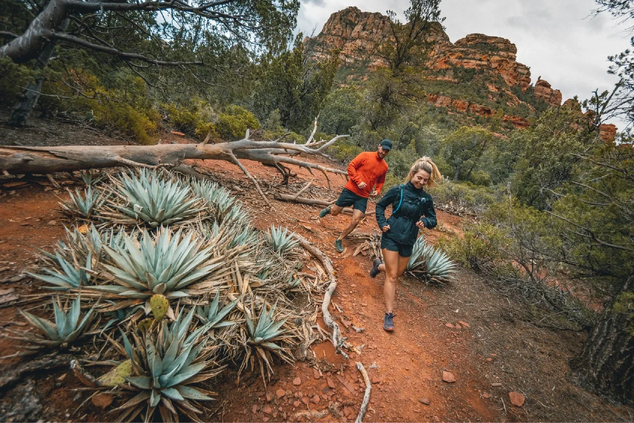 Two trail runners in long sleeves trail running in sedona. There is agave and green evergrenn shrubbery surrounded by red rock mountains.