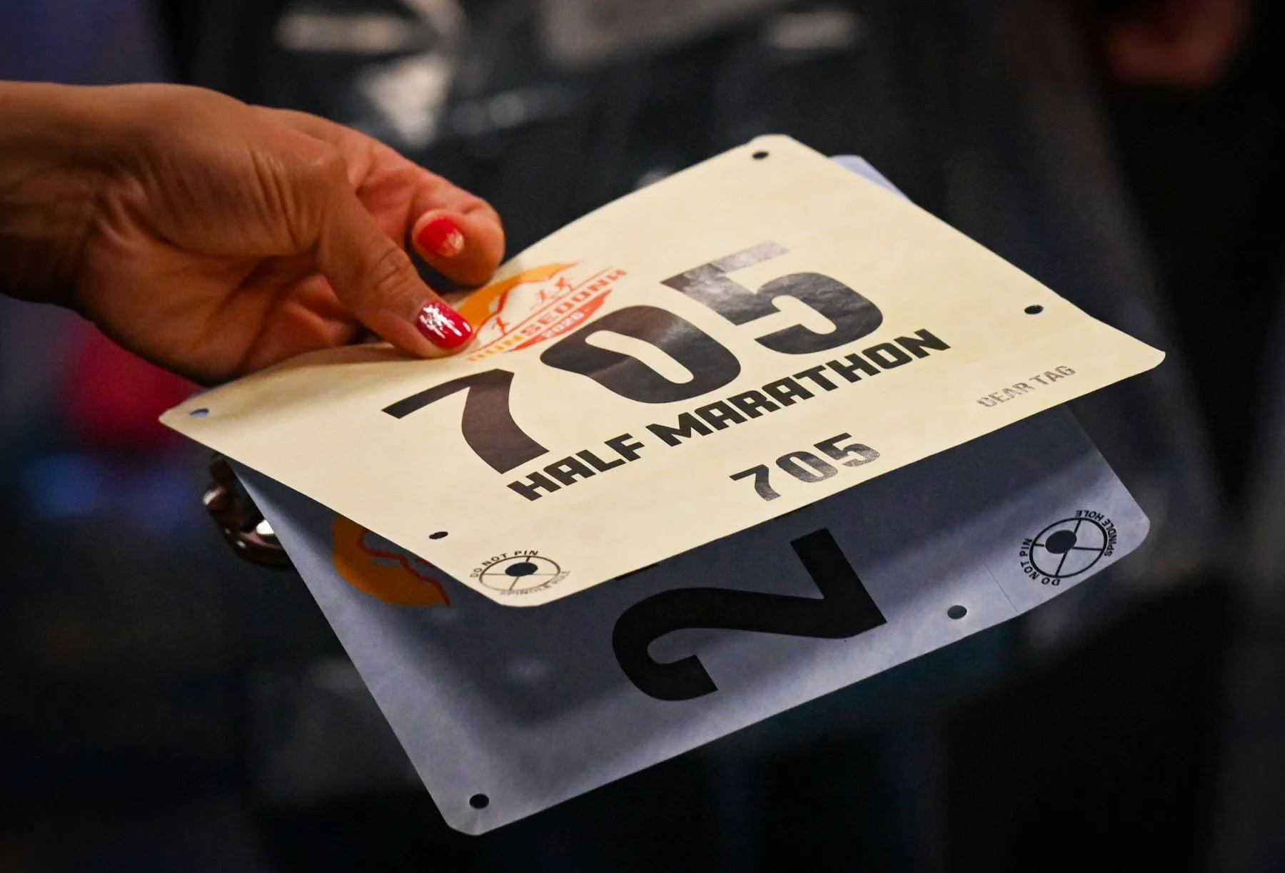 Close-up of a person's hand with red painted nails, holding a race bib with the number 705, labeled for a half marathon event.