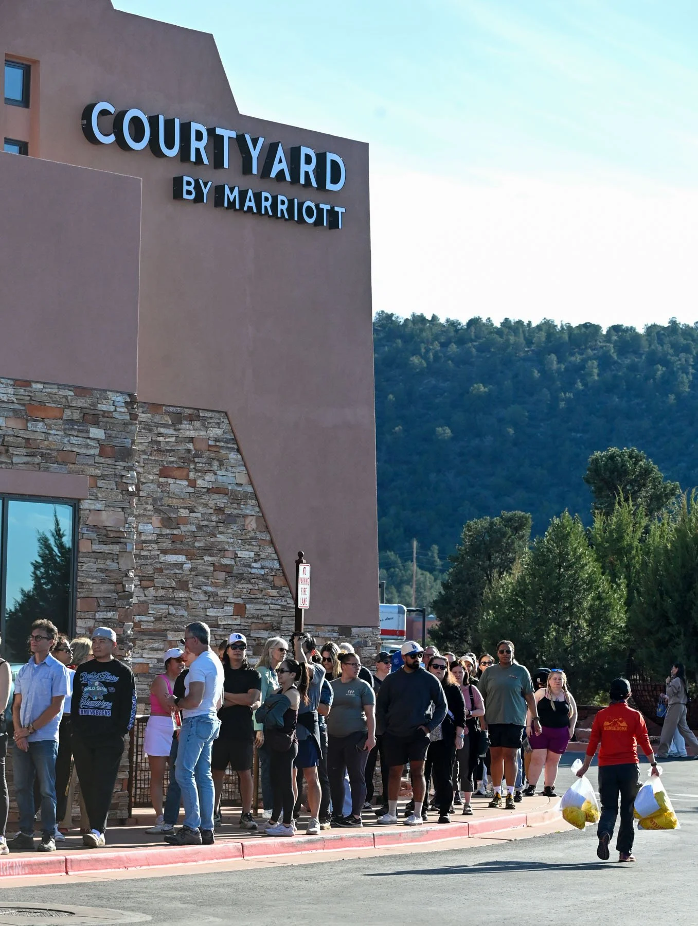 Line of people waiting outside a building labeled 'Courtyard by Marriott,' with mountains and trees in the background. Some people are wearing sunglasses and casual clothing, and a woman is carrying yellow bags.