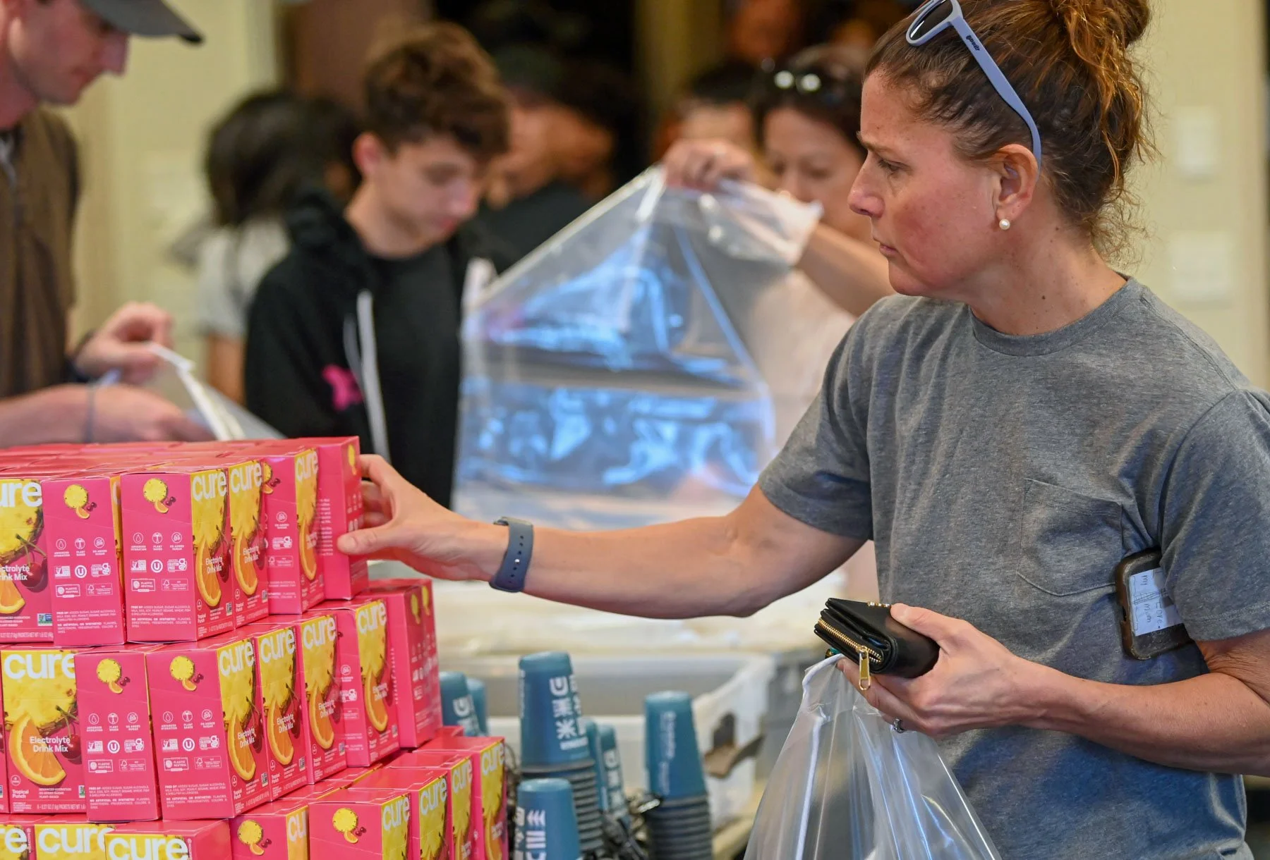 A woman shopping for electrolyte drink mix at a store, reaching for a pink box of Cure electrolyte drink mix with several other shoppers behind her.