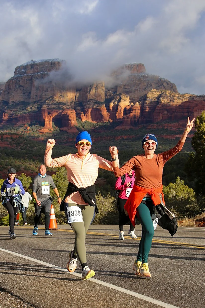 Two women running in a marathon, smiling and celebrating, with a mountainous landscape in the background.