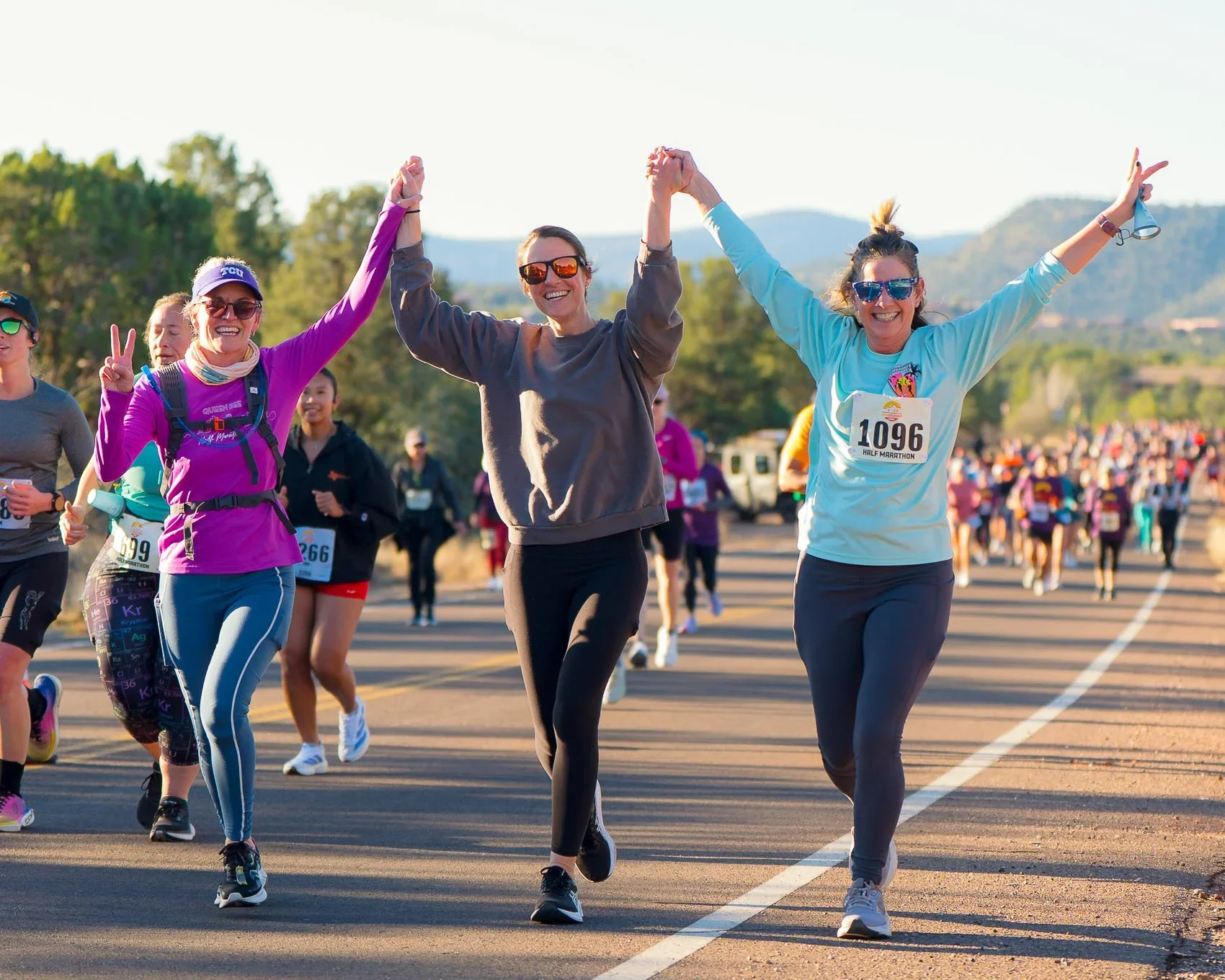 Three smiling women holding hands as they cross a marathon finish line, with scenic mountains behind them.