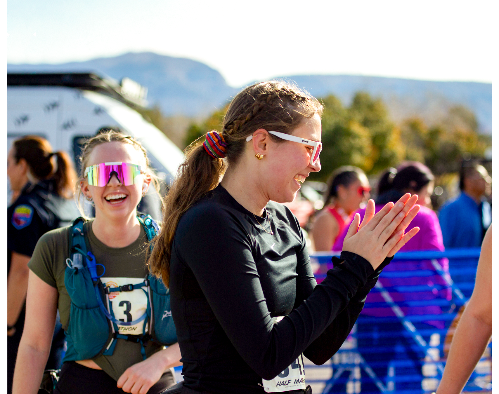 Two women participating in a half marathon or race, one smiling with sunglasses and the other clapping or cheering, with mountains and trees in the background.