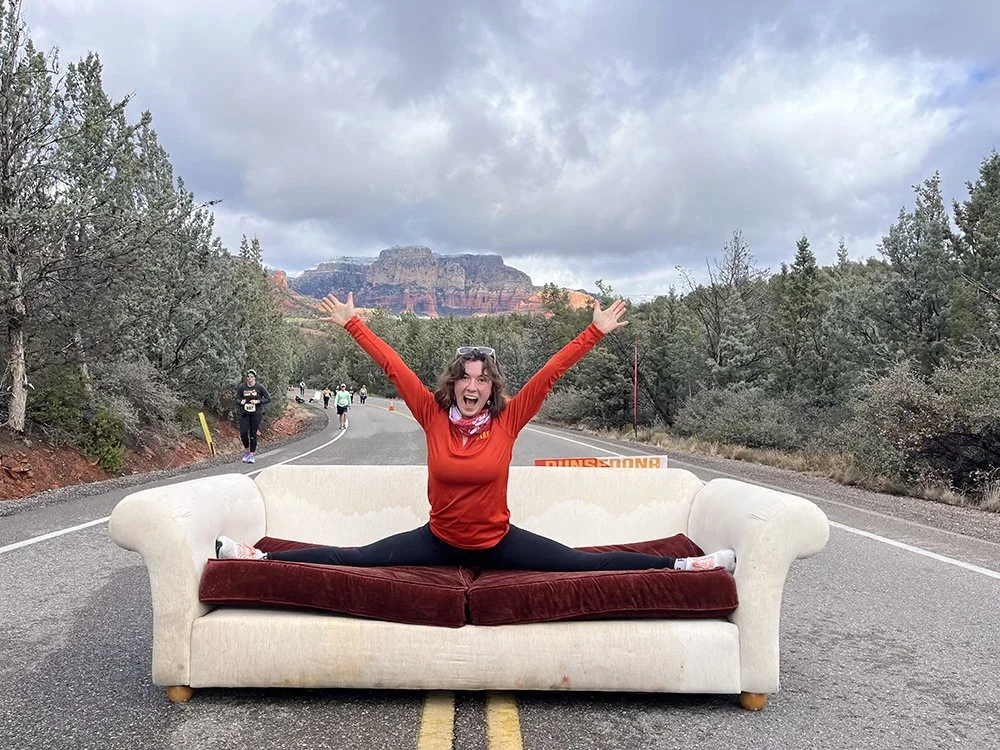 Road closures for RunSedona half marathon. The photo displays a runner in a splits across a couch in the middle of the road. She is wearing a bright red loing sleeve, the couch has red cushions and a beige body.