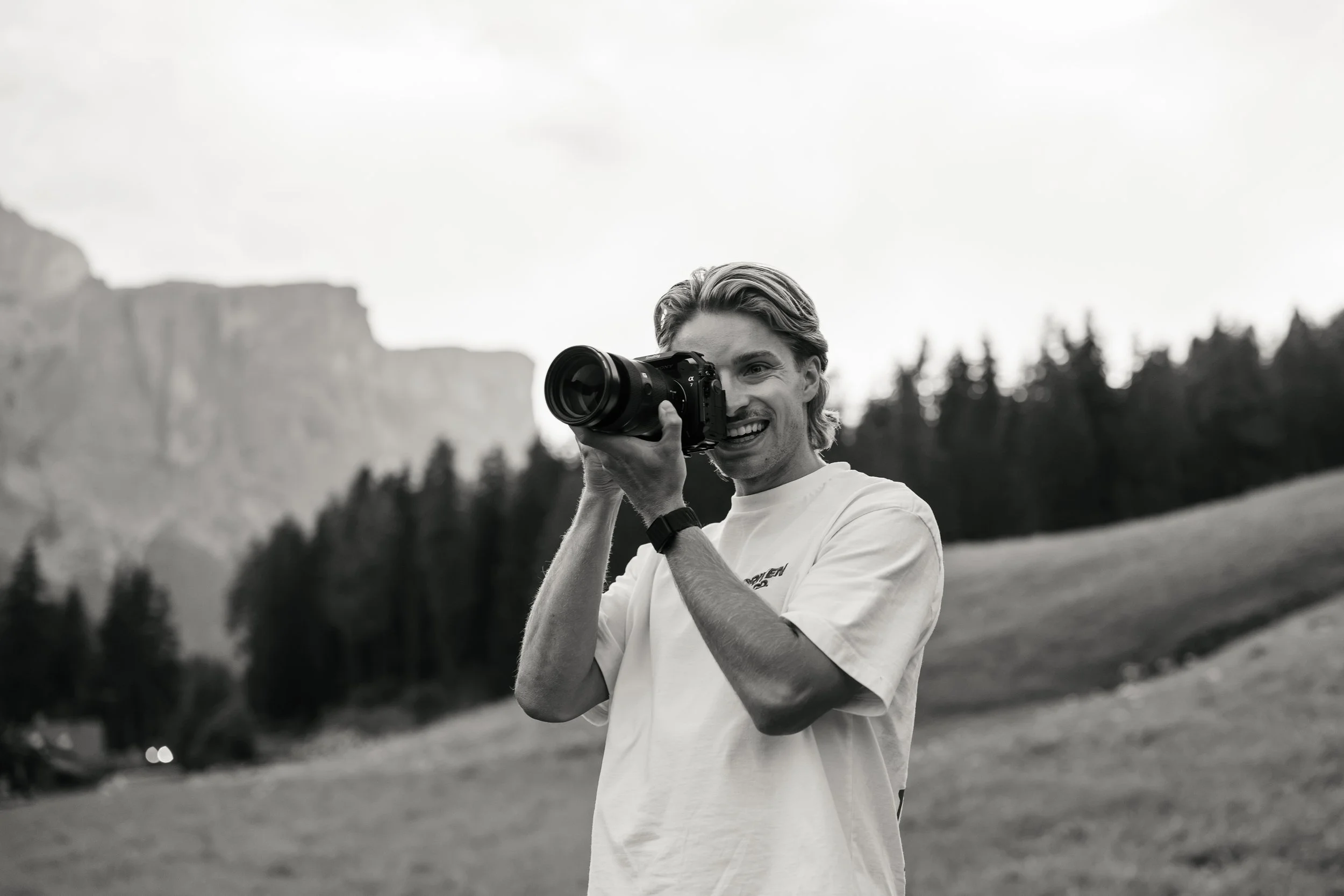 Ein junger Mann mit freundlichem Gesicht hält eine Kamera hoch, um ein Foto zu machen, im Freien mit Bäumen und Bergen im Hintergrund.