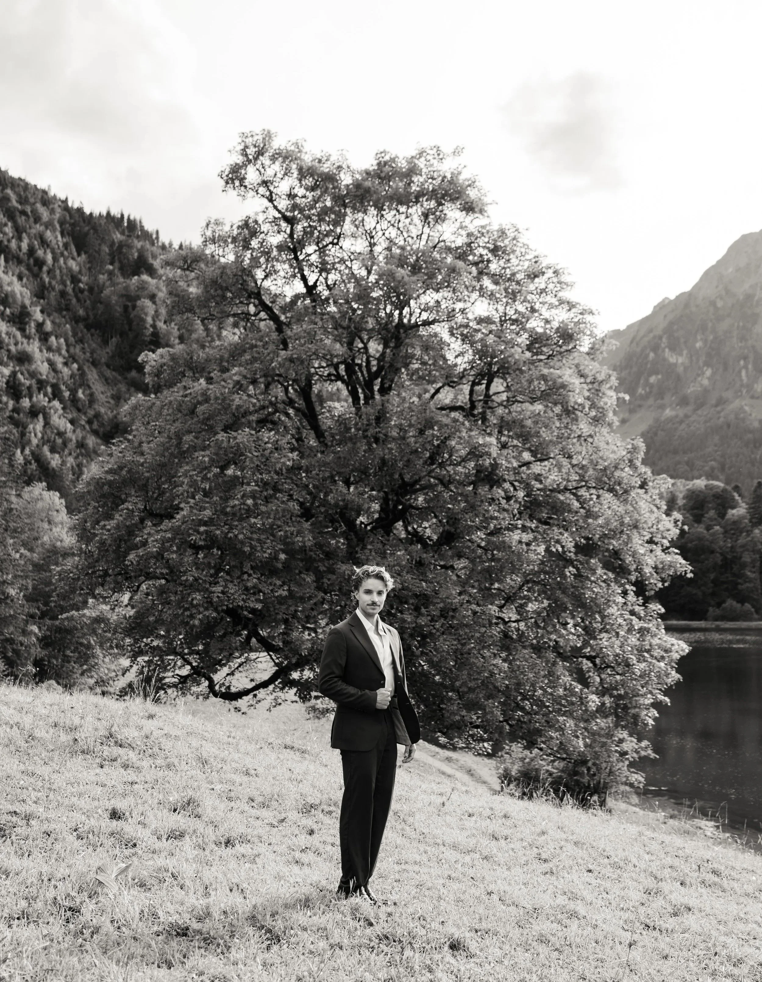 Schwarz-weiß-Foto eines Mannes im Anzug, der an einem Fluss steht, im Hintergrund ein großer Baum und Berge.