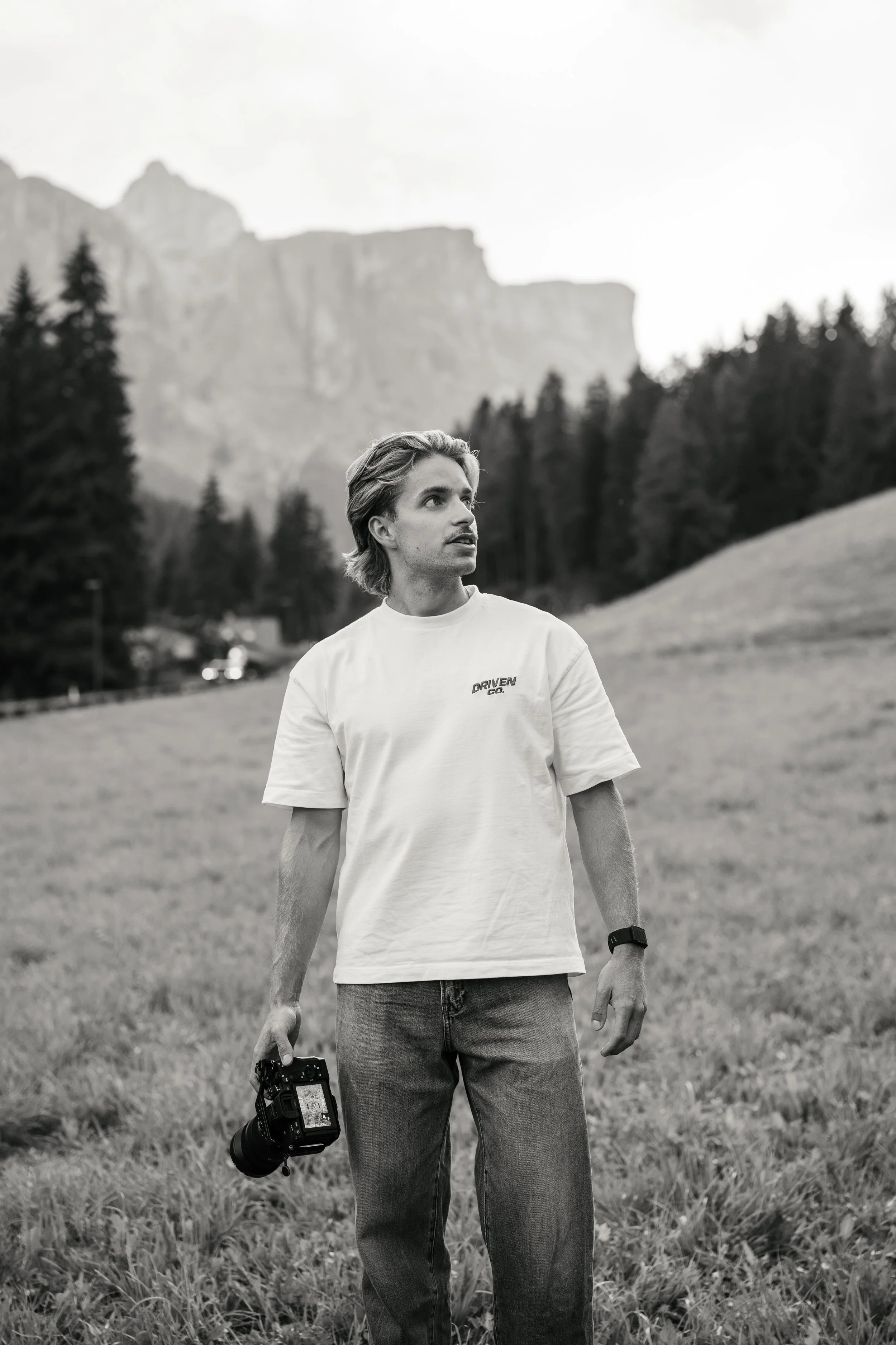 Junger Mann mit Kamera in der Hand, trägt T-Shirt mit Aufdruck, steht in einer Wiese vor Bergen und Bäumen, schwarzen und weißen Fotostil.