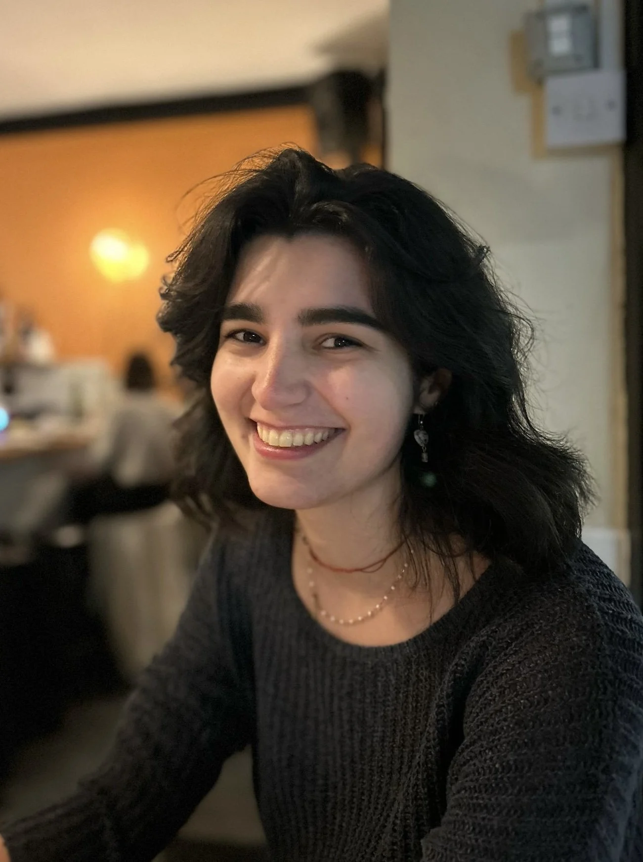 A young woman with dark, wavy hair and white teeth smiling in a dimly lit indoor setting, wearing earrings, a necklace, and a dark sweater.