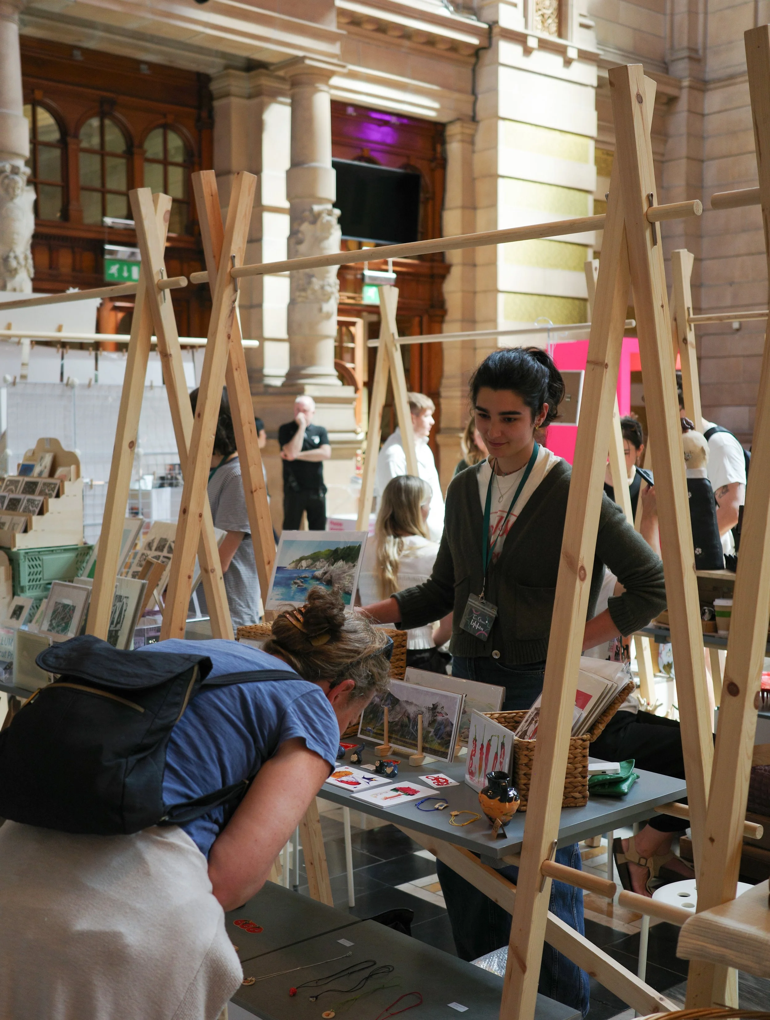 People at a craft or jewelry-making event in a spacious, ornate hall with high ceilings and classical columns. They are working on handmade jewelry or small art projects at tables.