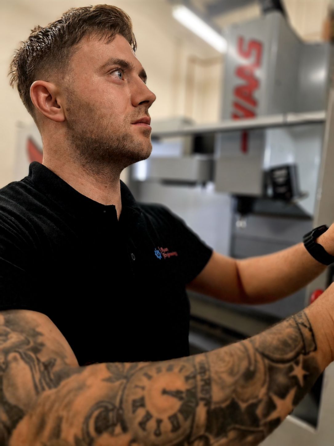 Side profile of a man with short hair, wearing a black shirt, standing in front of a machine, with another person’s arm and tattooed forearm visible in the foreground.