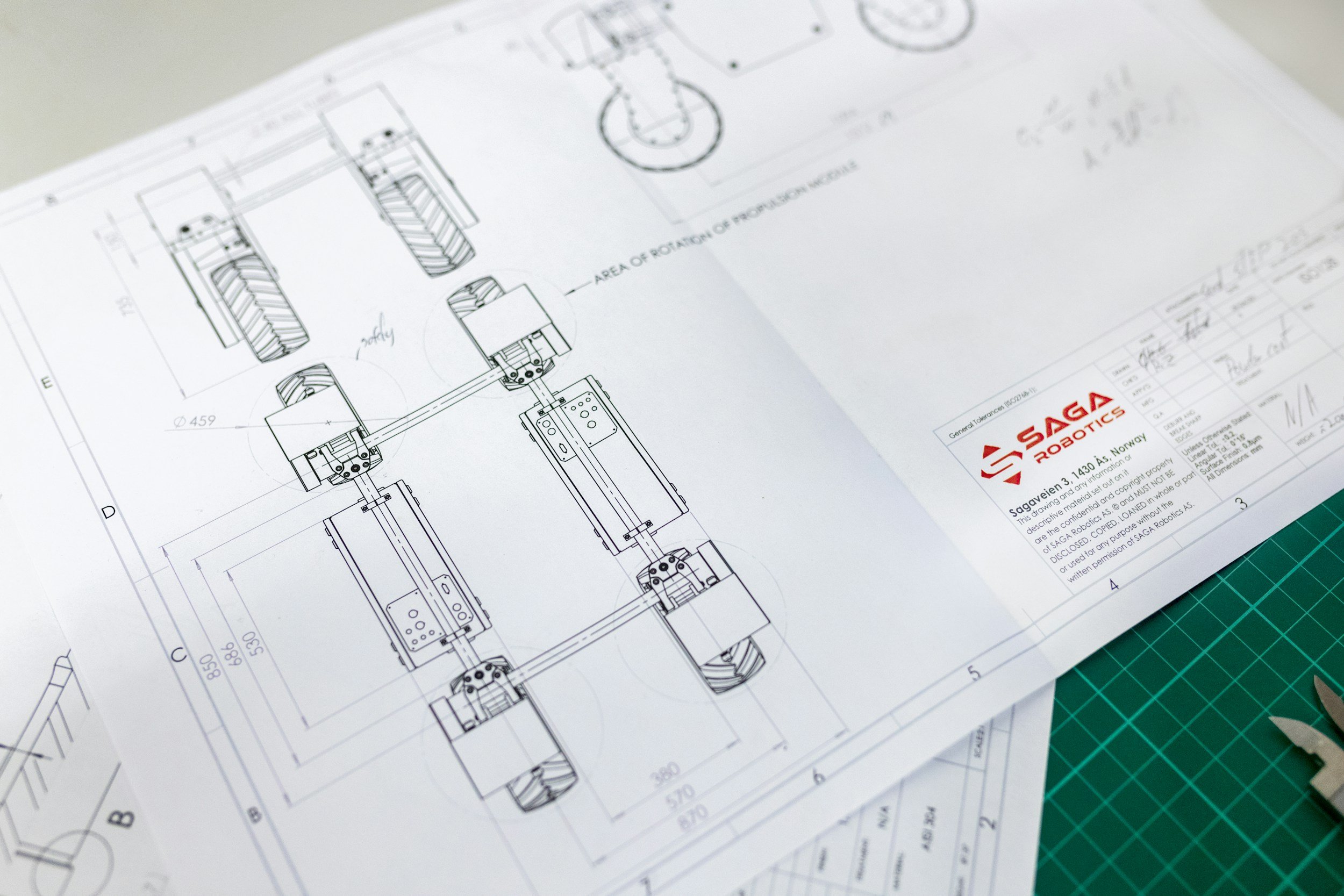 Technical blueprint or engineering drawing of a robotic system on white paper with the logo and address of Saga Robotics in Norway, placed on a green cutting mat.