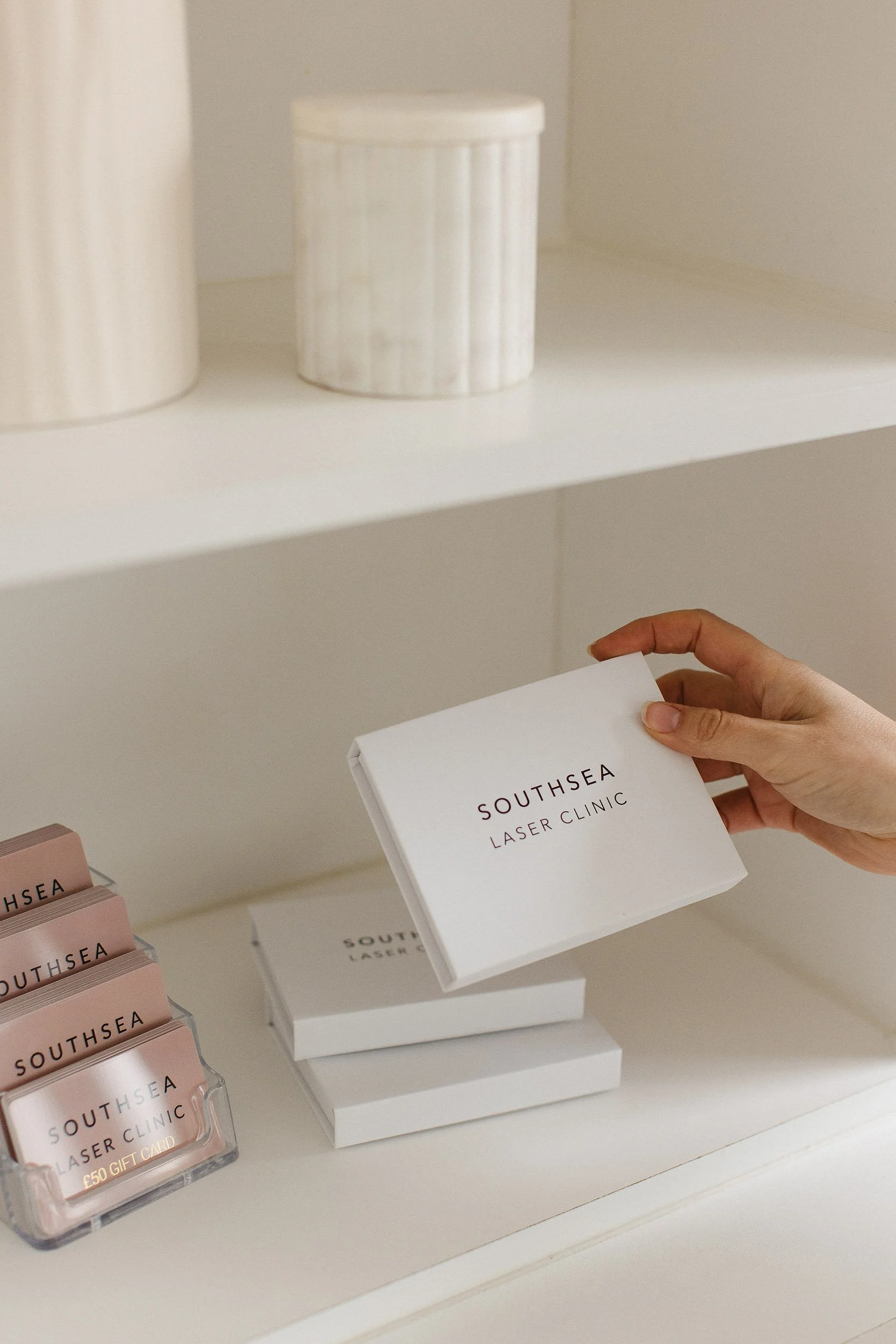 Hand holding a white box labeled 'Southsea Laser Clinic' on a shelf with other similar boxes and pink gift cards, interior of a modern spa or clinic.