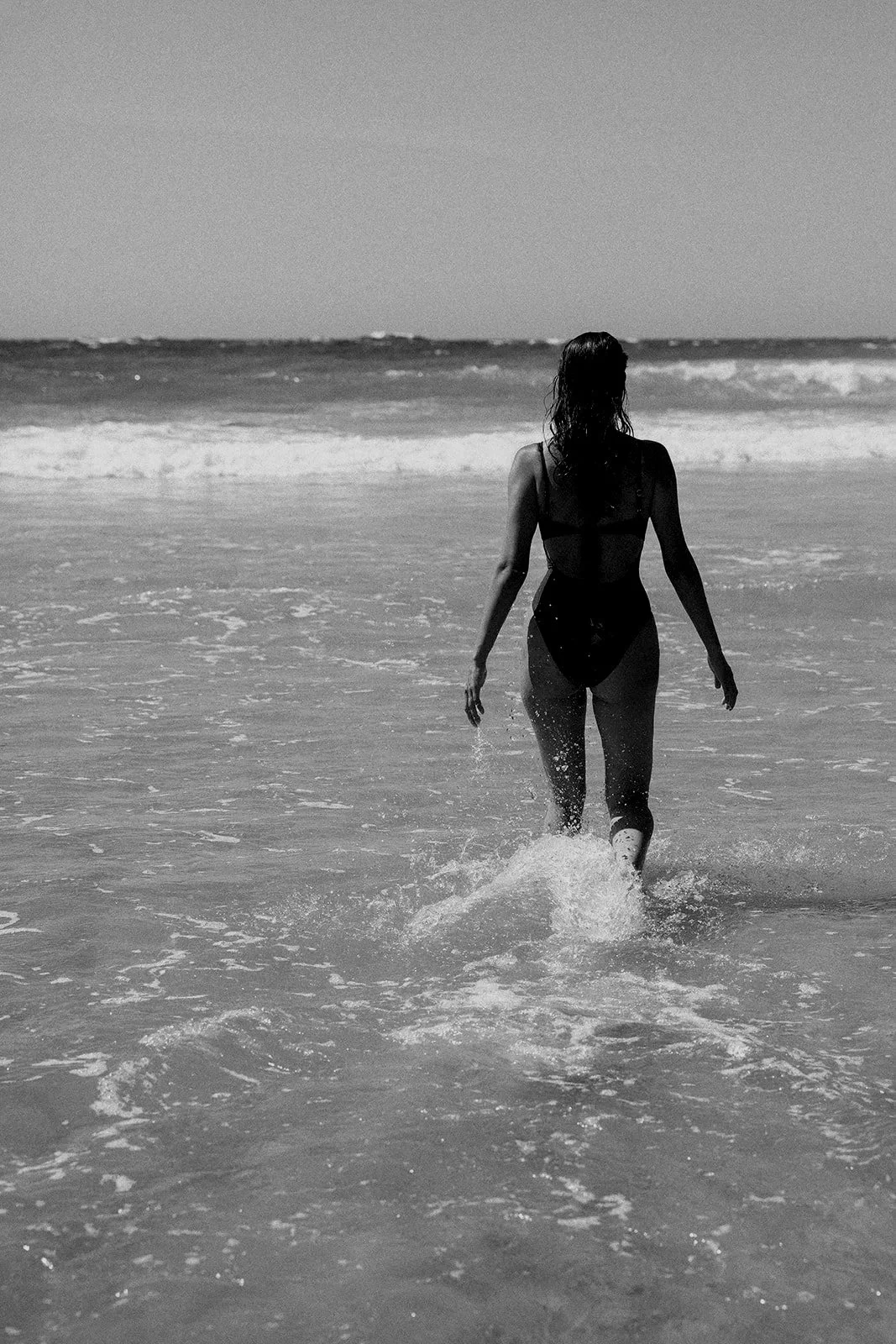 A woman wading into the ocean, viewed from behind, with waves and the horizon in the background, black and white photo.