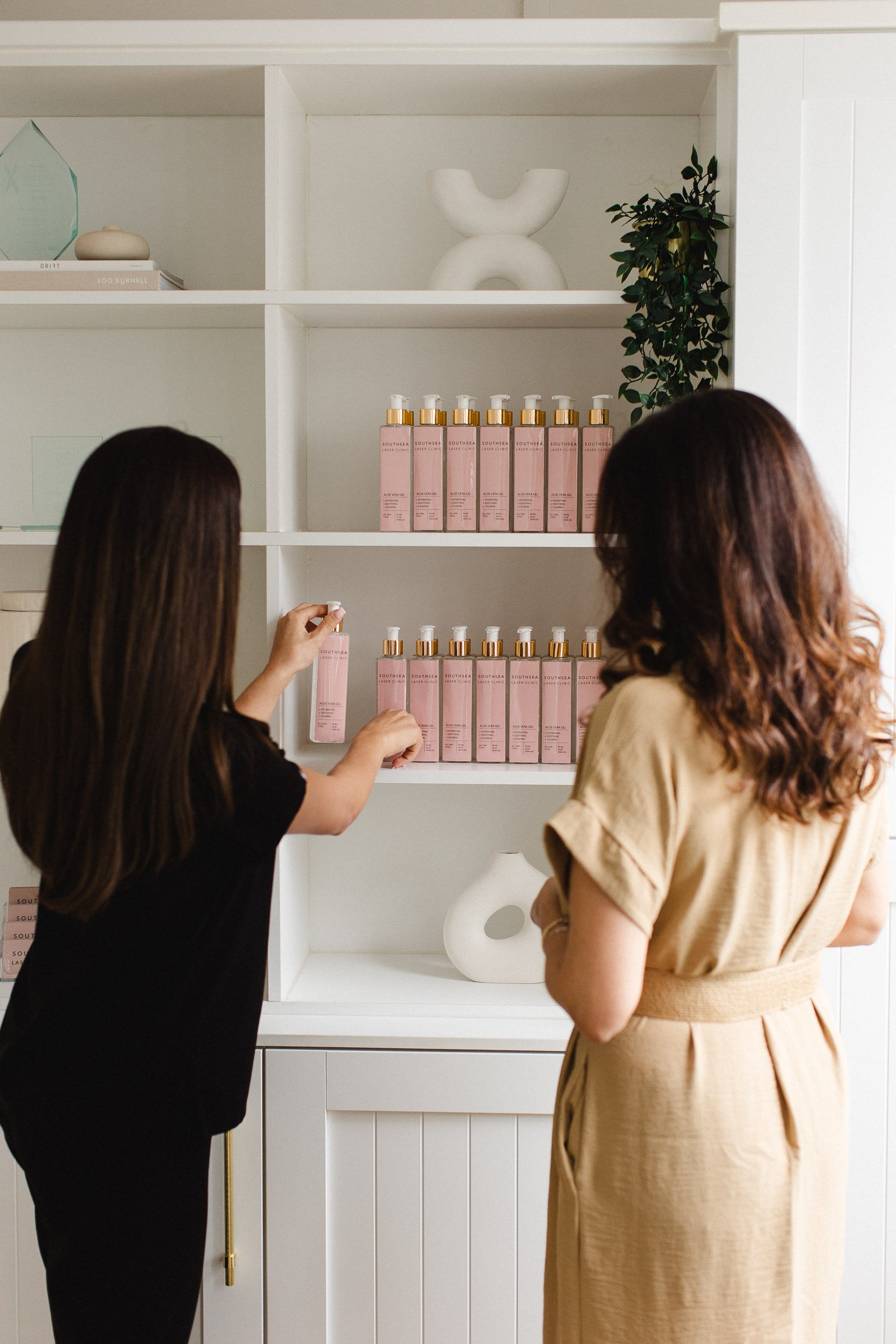 Two women looking at shelves of skincare products with pink bottles in a minimalist white room.