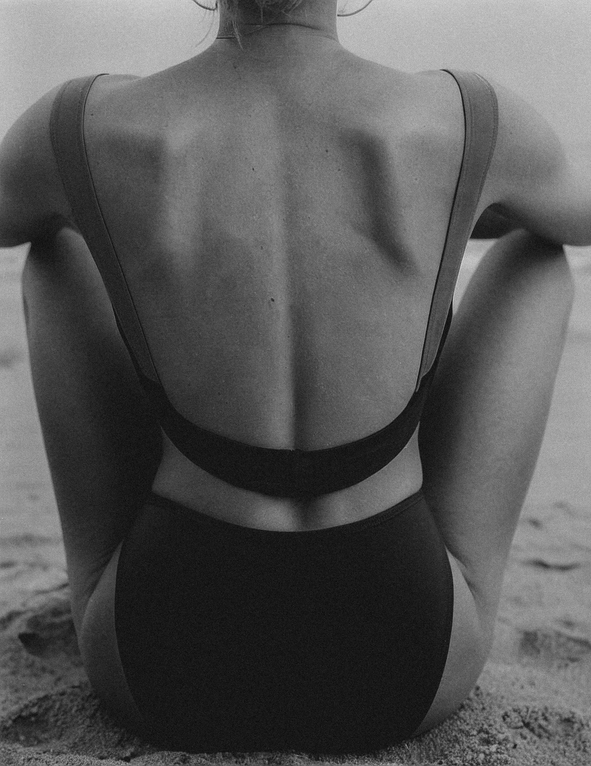 A person sitting on the sand with their back facing the camera, wearing a black top and black bottom, at the beach.