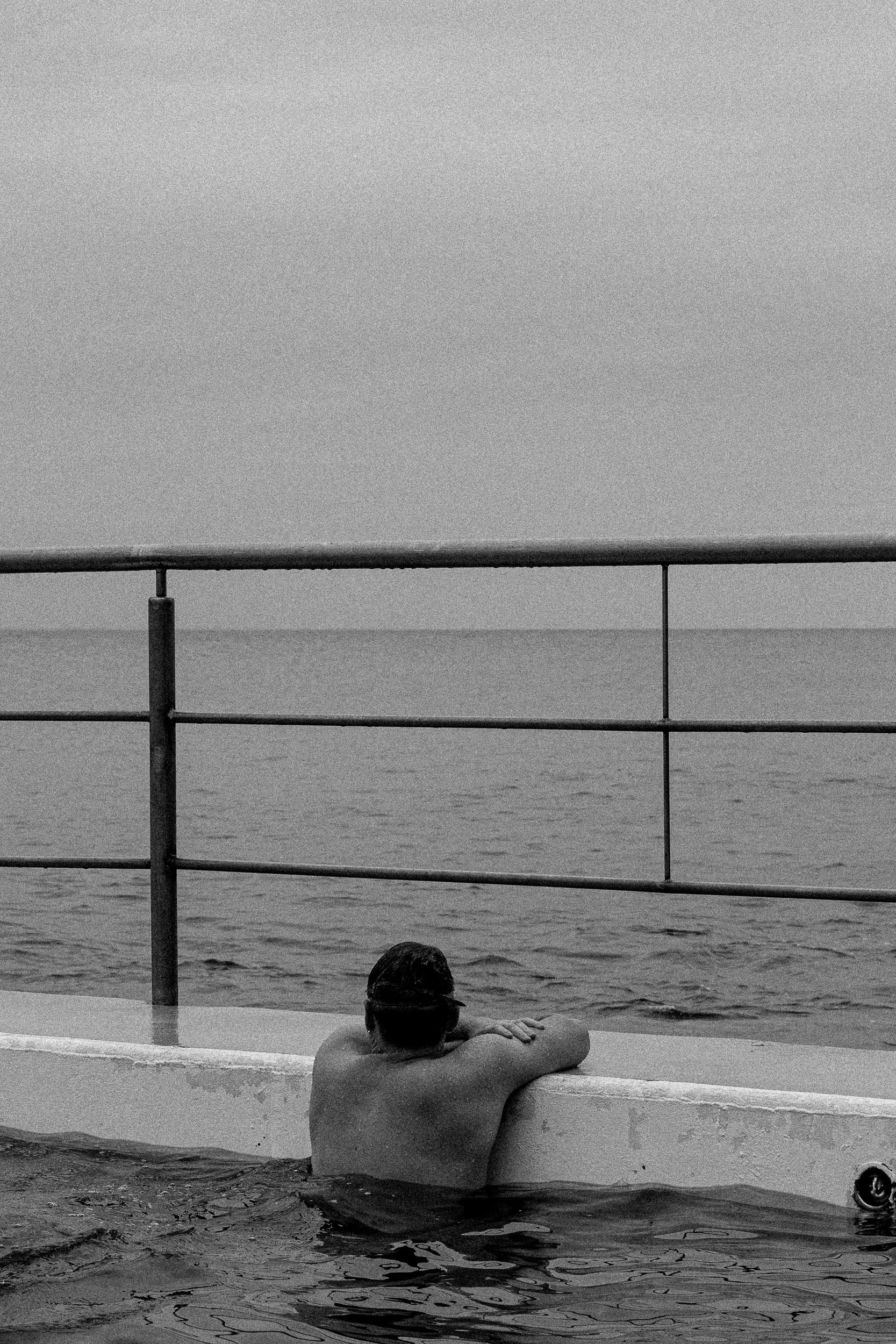 A man leaning on a concrete edge at a swimming pool, looking out at a body of water beyond a metal railing.