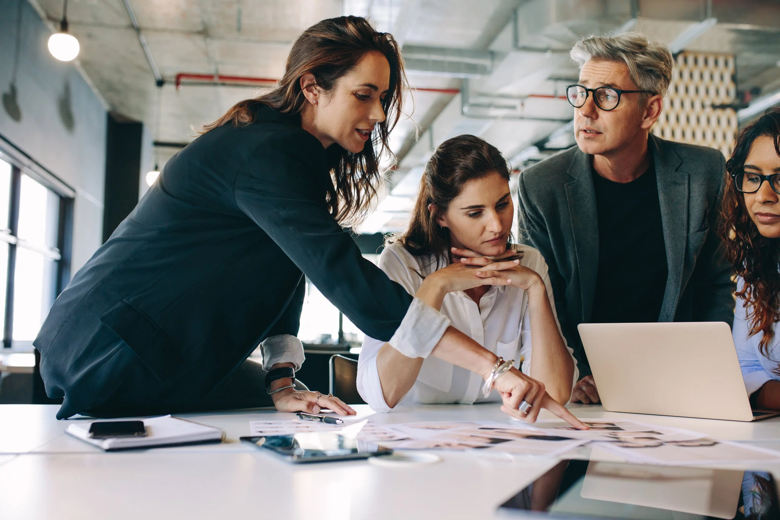 Vier Menschen bei einem Meeting in einem modernen Büro, einer erklärt etwas auf einem Tisch mit Papieren, Laptop und Tablets.