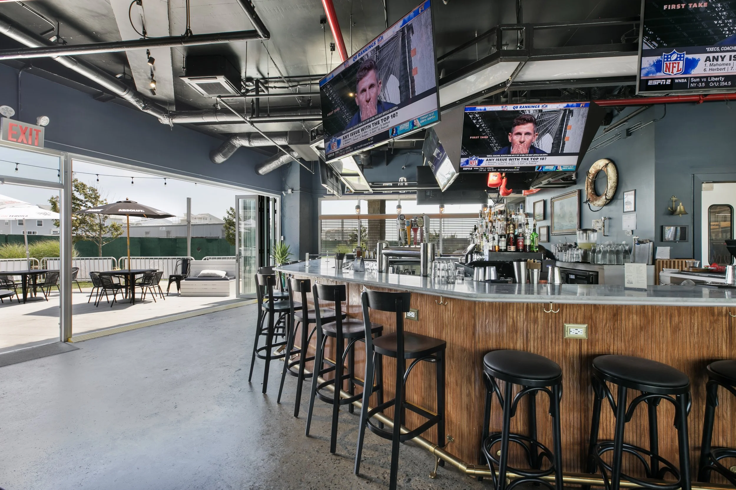 Interior of a bar or restaurant with a marble countertop, barstools, and multiple television screens hanging from the ceiling. Outside patio area with tables, chairs, and umbrellas visible through glass doors and windows.