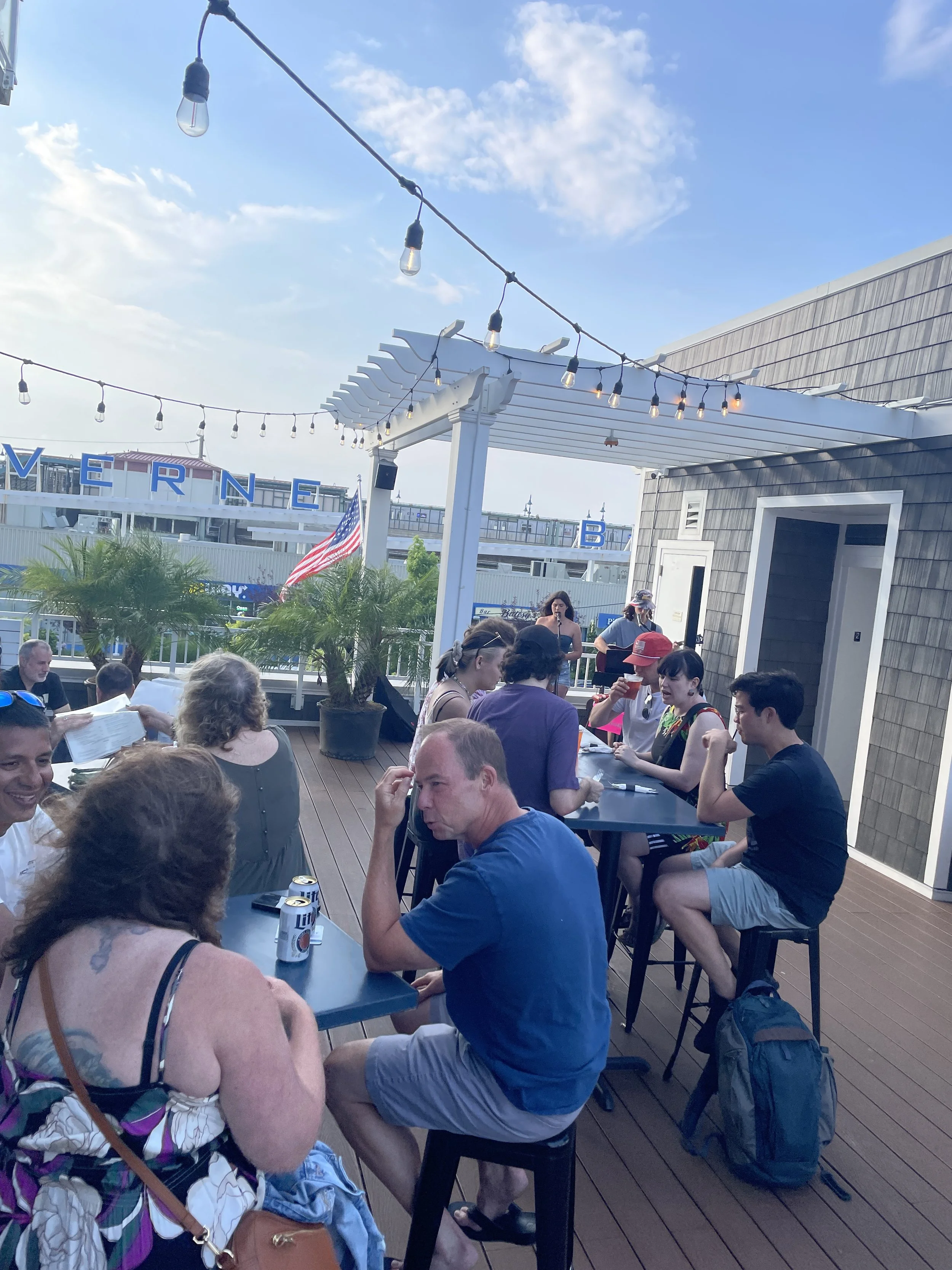 People enjoying outdoor dining on a patio under string lights with a clear blue sky and some clouds, with a sign reading 'VERE' visible in the background.