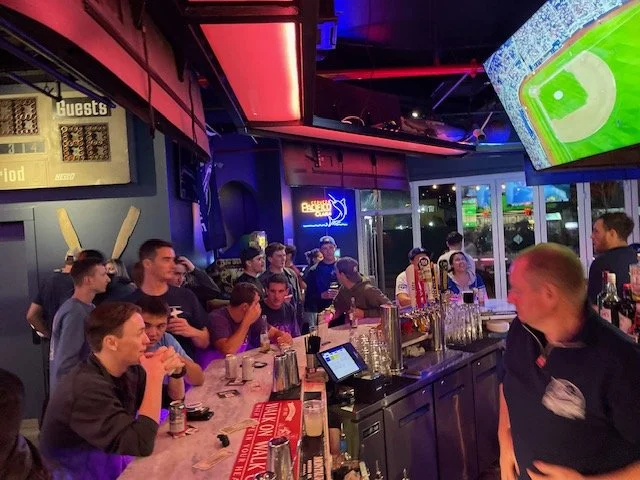 Group of young adults sitting at a sports bar watching a game on multiple TVs, with a bartender behind the counter and neon signs on the wall.