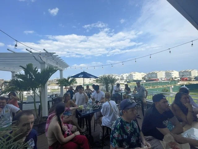 People dining outdoors on a patio with string lights, potted palms, and a view of buildings and a waterway in the background.