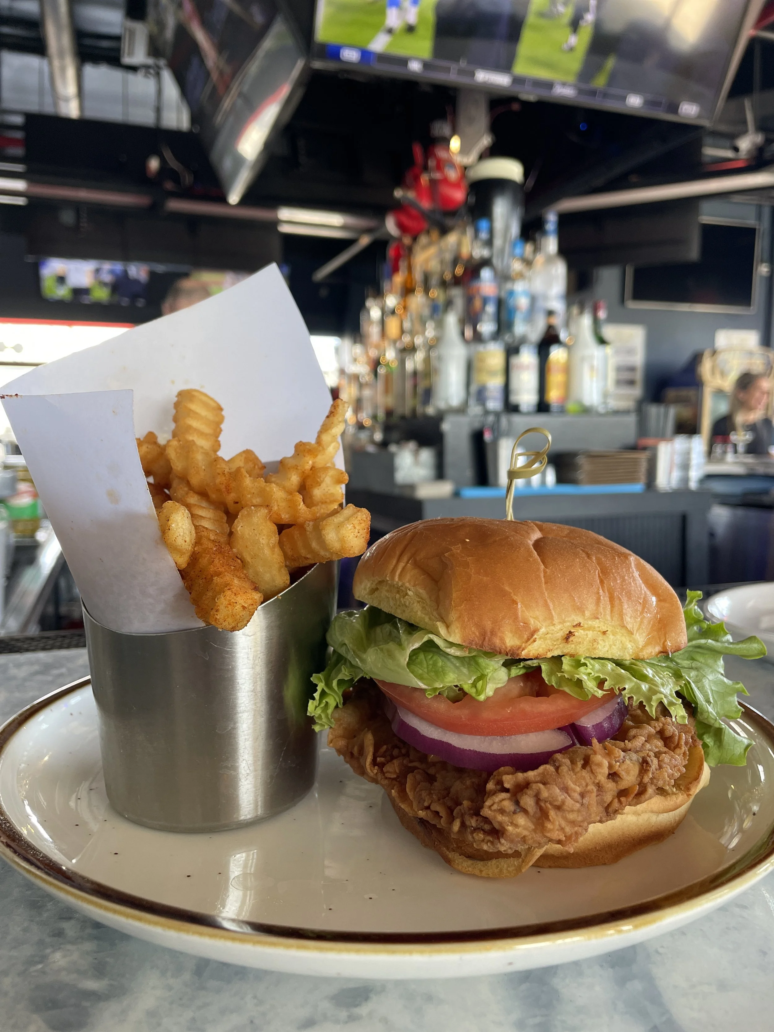 A plate with a fried chicken sandwich topped with lettuce, tomato, onion, and pickles, along with a side of crinkle-cut French fries in a small metal container.