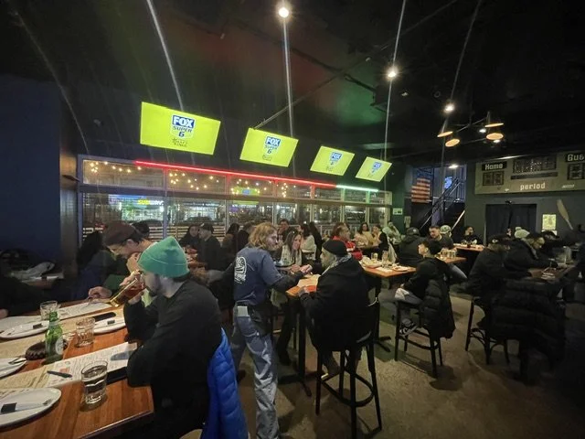People dining and watching TV screens in a restaurant or bar setting with illuminated signs and dark interior.
